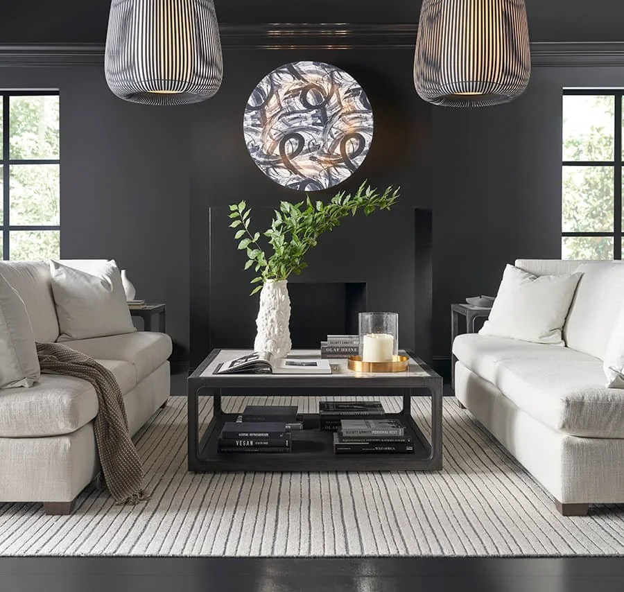 Living room with black wall, two white sofas, striped area rug, black coffee table with books, a vase with greenery, candles, and a decorative wall art piece, illuminated by black and white pendant lights, with windows letting in natural light.