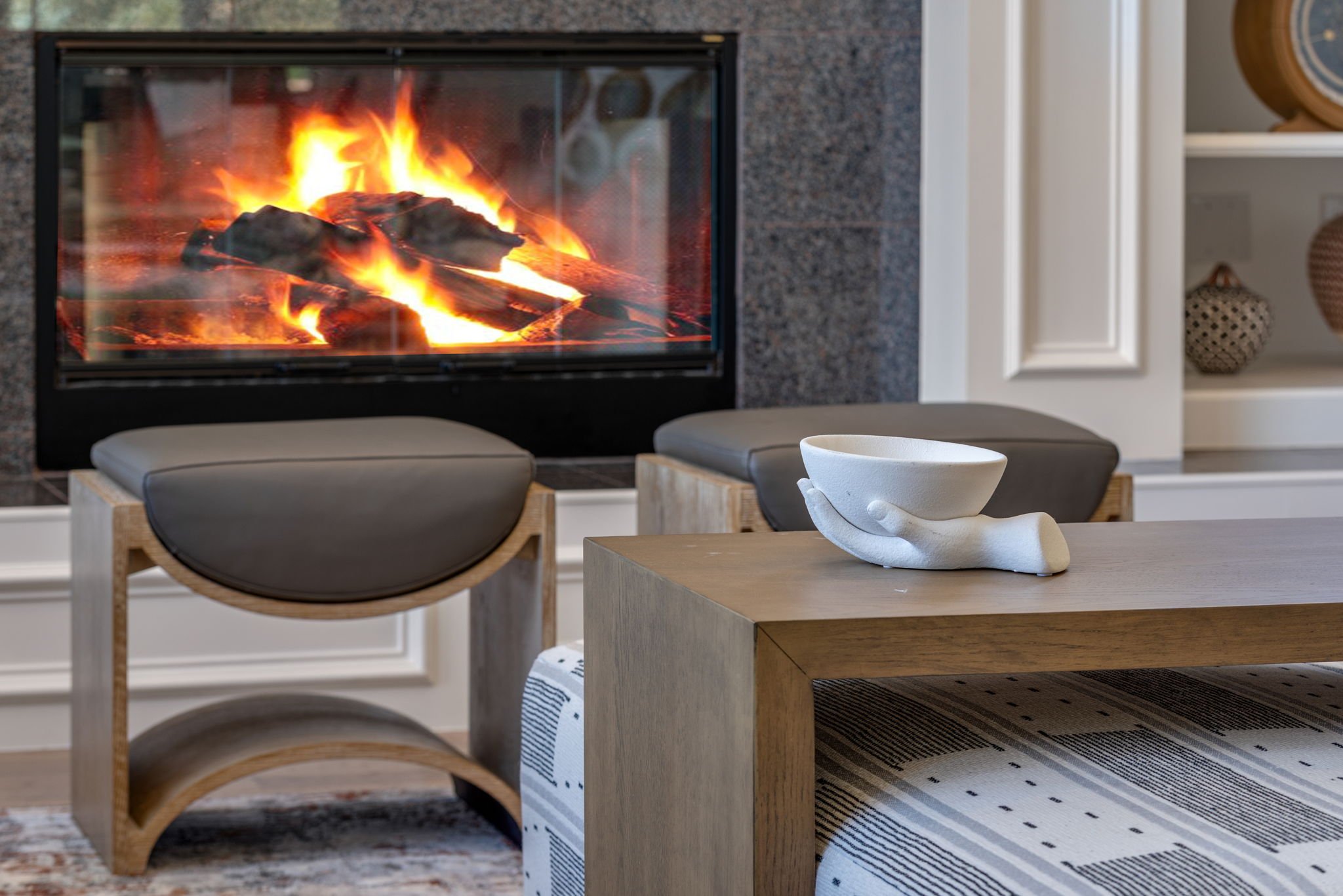 Living room with a modern fireplace, two chairs with wooden frames and dark cushions, and a wooden coffee table with a white ceramic hand holding a bowl.