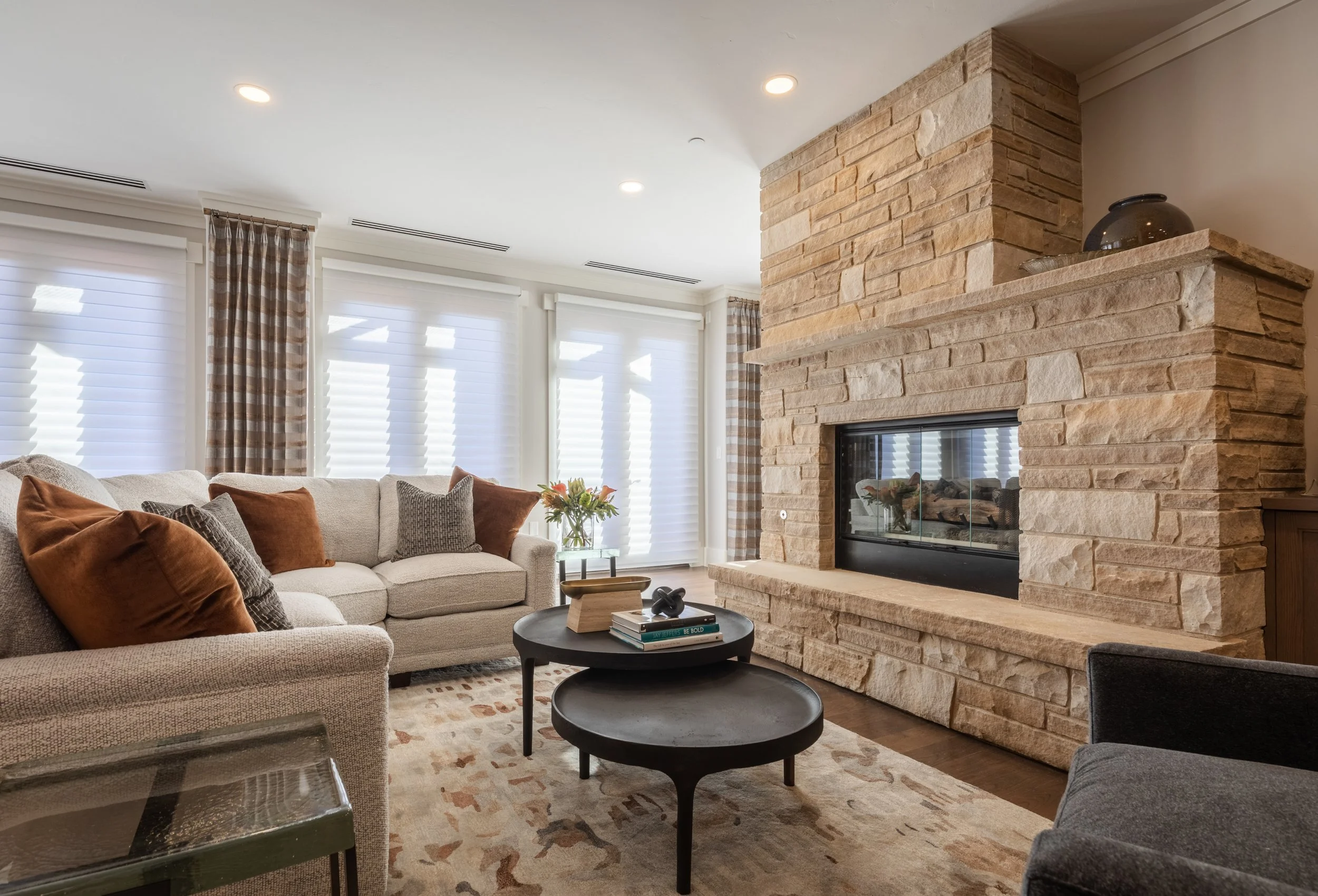 Living room with beige sofa, pillows, fireplace with stone mantel, large windows with white blinds and plaid curtains, coffee tables, and decorative items.