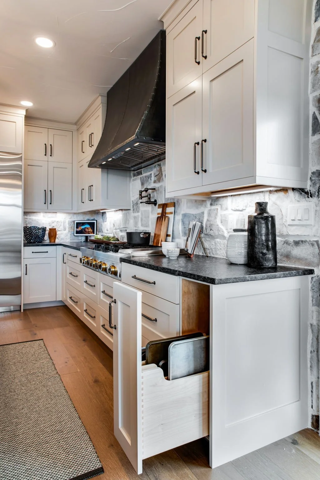Modern kitchen with white cabinets, black handles, a black range hood, and a stone backsplash. A pull-out drawer contains baking sheets, and countertop decor includes vases and small bowls.