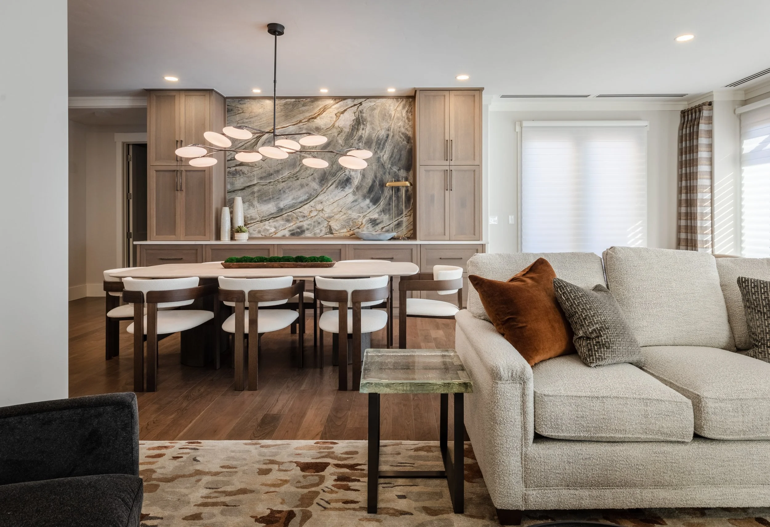Living room with beige sofa, pillows, side table, dining area with wooden table, chairs, and chandelier, built-in cabinets, marble backsplash, and windows with blinds and curtains.