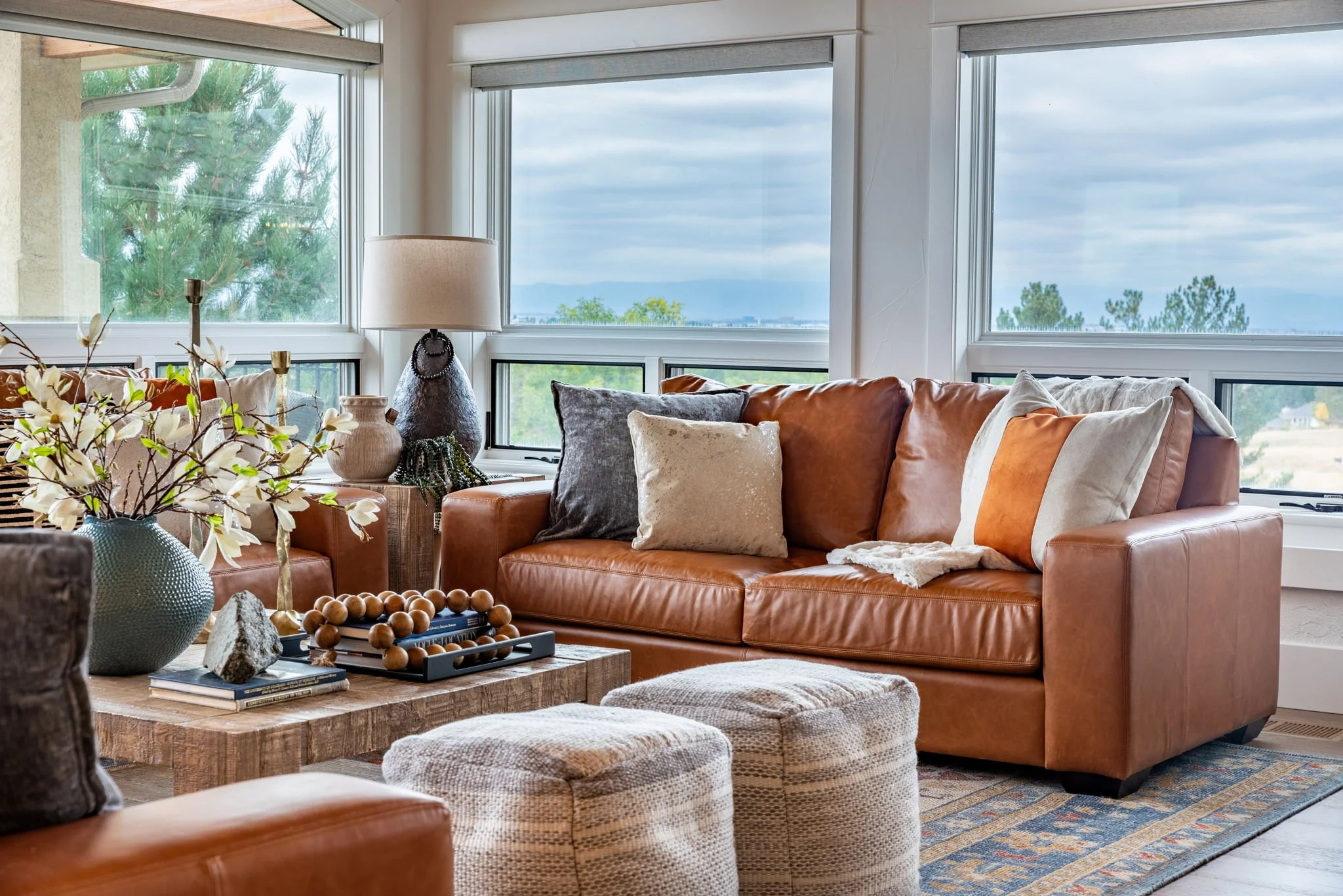 Living room with a brown leather sofa decorated with various pillows near large windows showing an outdoor view of trees and cloudy sky.
