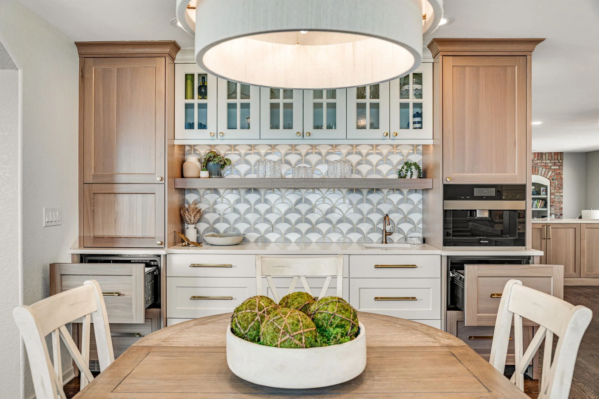 Kitchen with light wood and white cabinets, patterned tile backsplash, and a round dining table with a bowl of decorative green moss balls