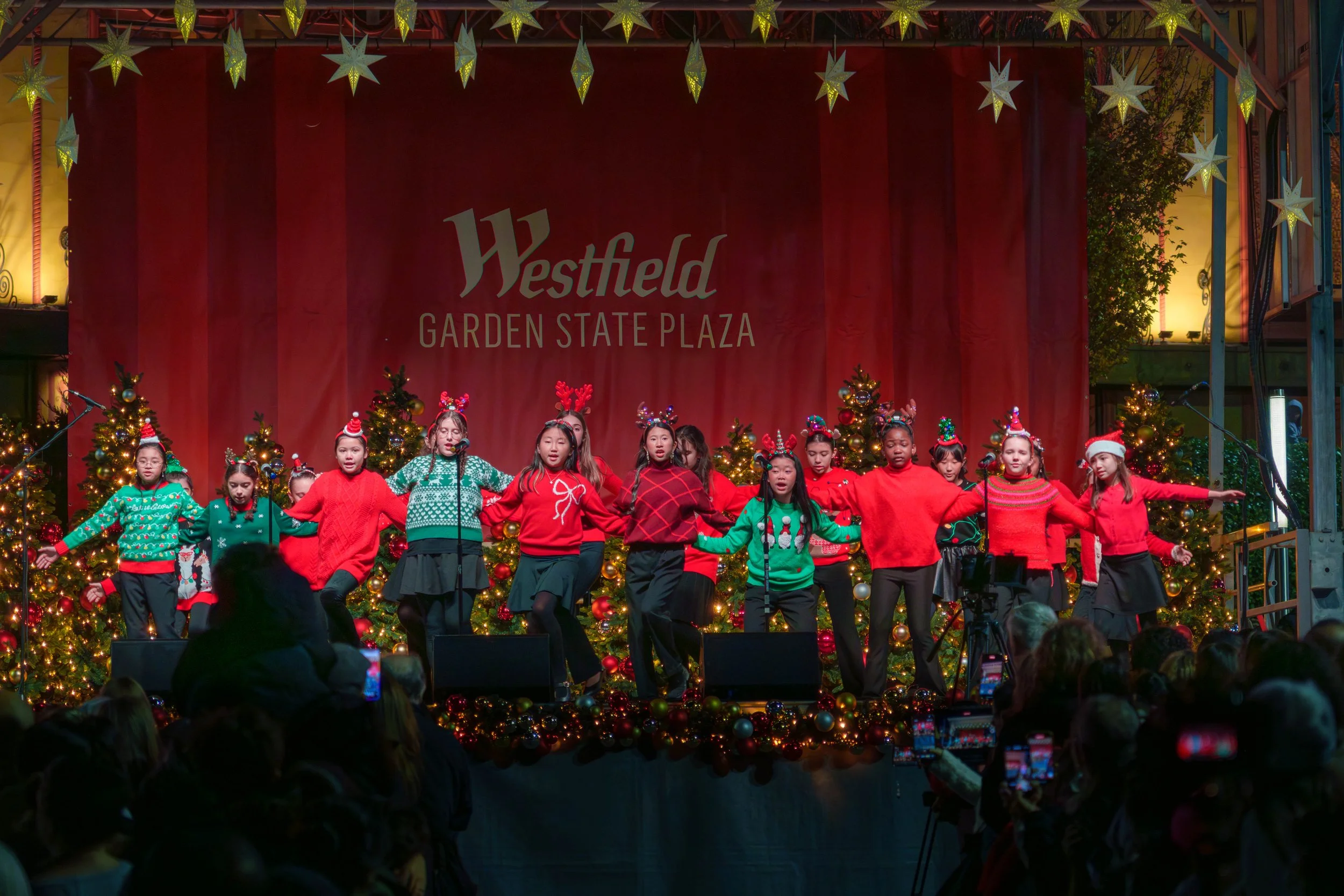 Young children performing a holiday dance on stage at Westfield Garden State Plaza, decorated with Christmas trees, ornaments, and star-shaped lights.