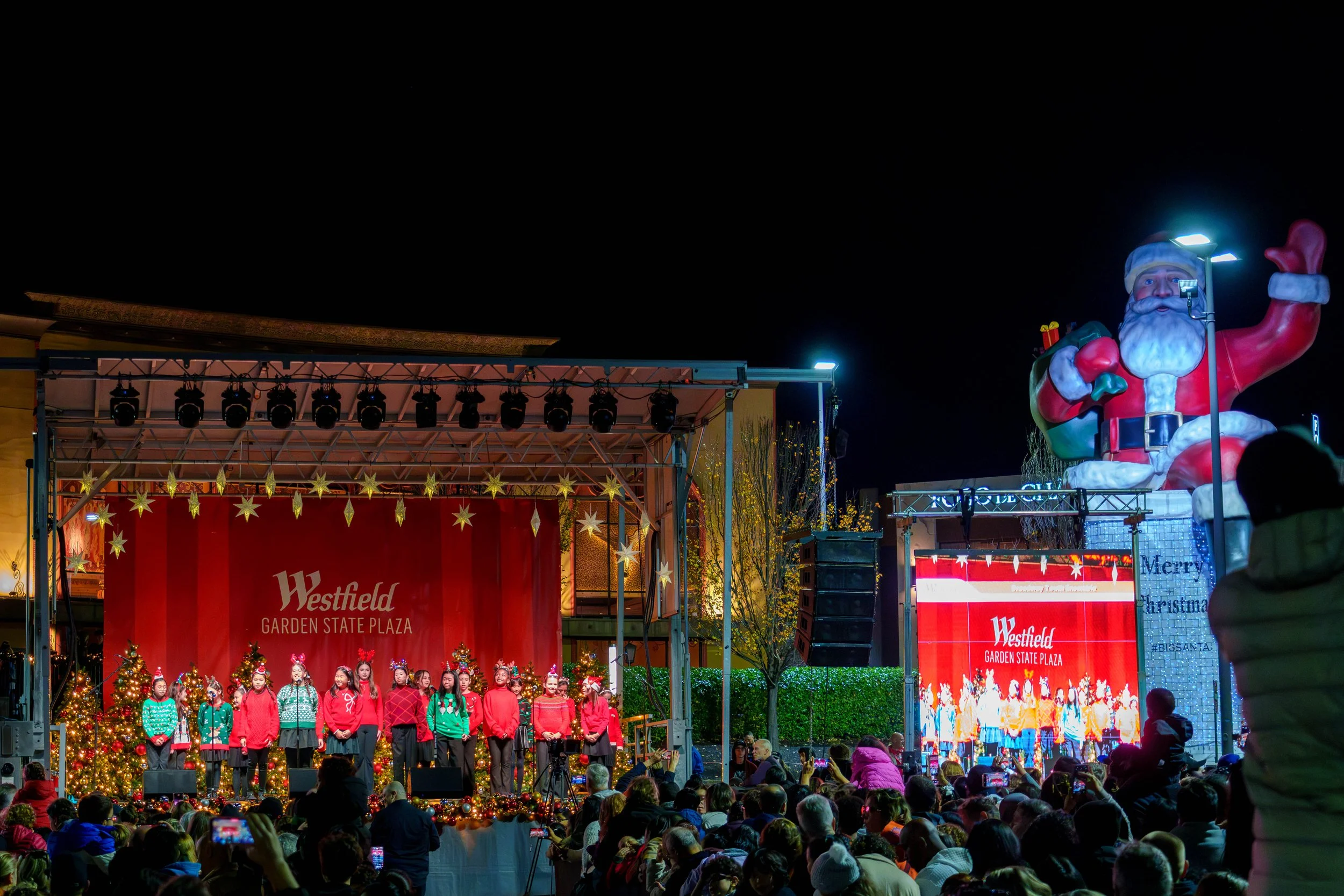 Group of children performing on stage at Westfield Garden State Plaza Christmas event, decorated with Christmas trees and lights, with a large inflatable Santa Claus nearby