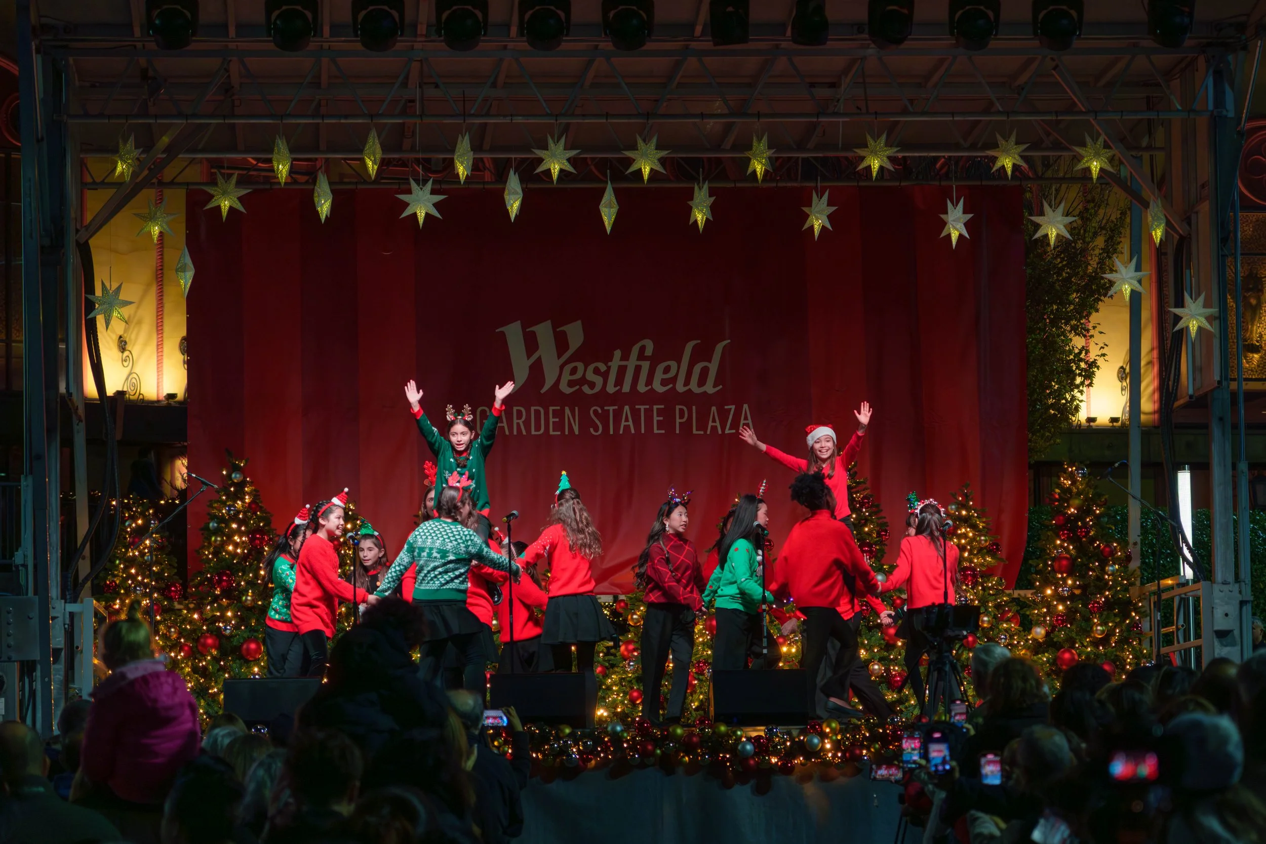 Children performing on a Christmas-themed stage at Westfield Garden State Plaza, decorated with Christmas trees and lights, with some children wearing Christmas sweaters and Santa hats.