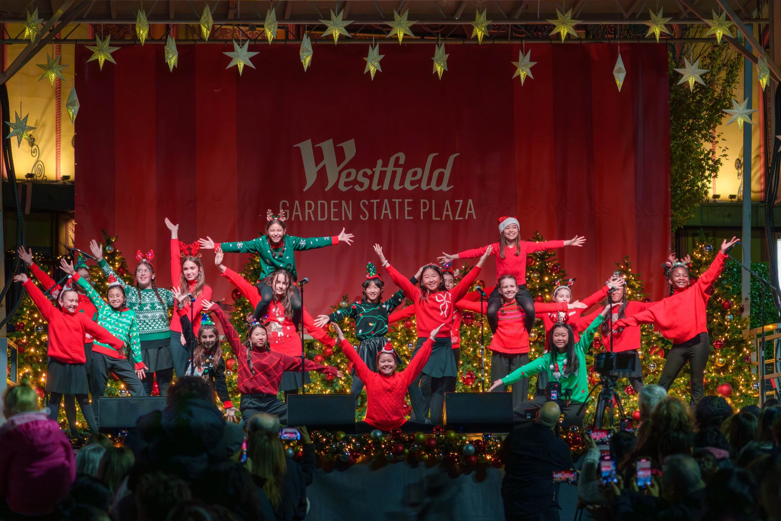 Children performing a Christmas dance on stage at Westfield Garden State Plaza, decorated with Christmas trees, ornaments, and festive lights, with an audience watching.