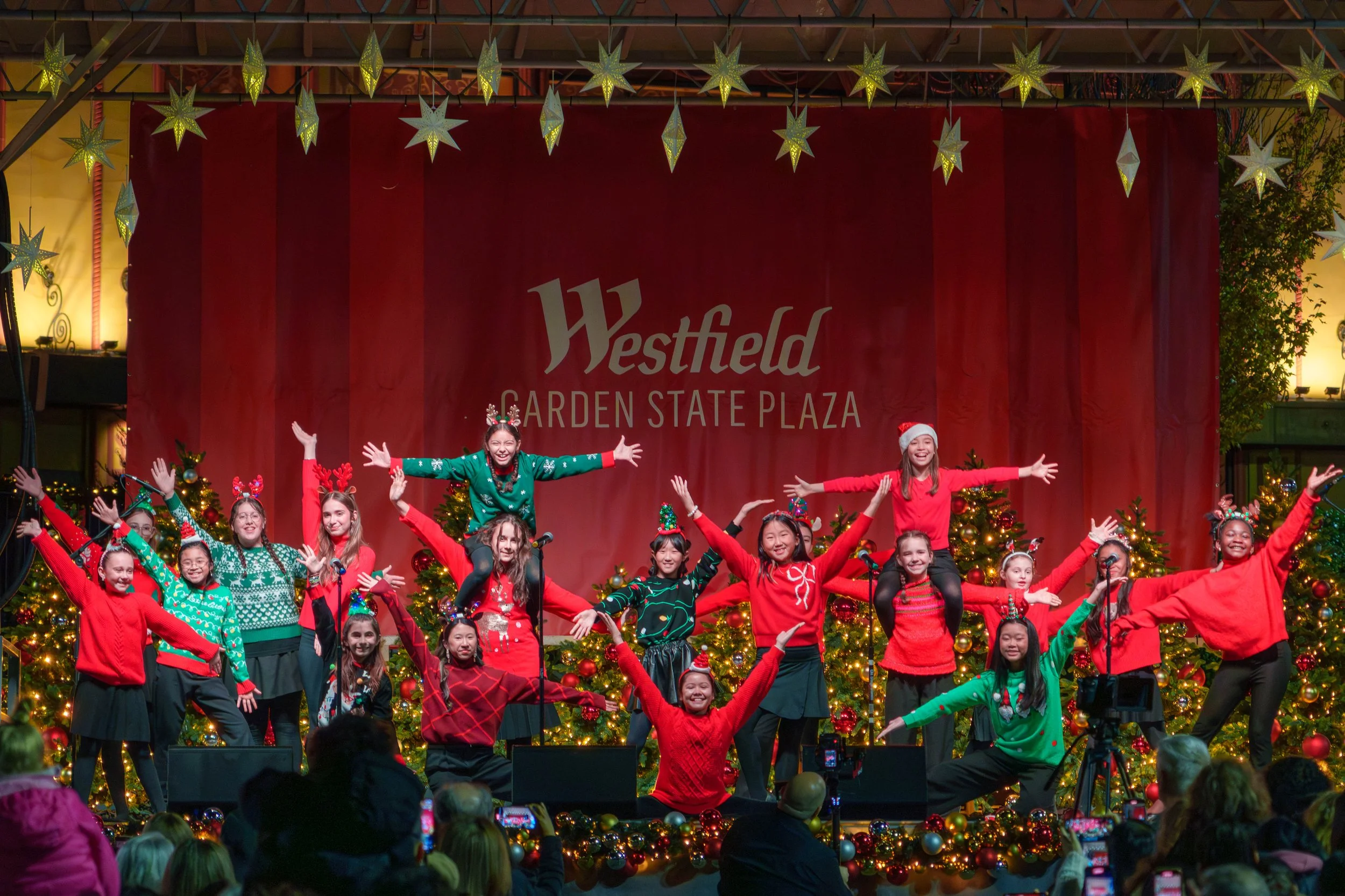 A group of children performing a Christmas-themed dance on stage at Westfield Garden State Plaza, decorated with Christmas trees and holiday lights, during a festive event.
