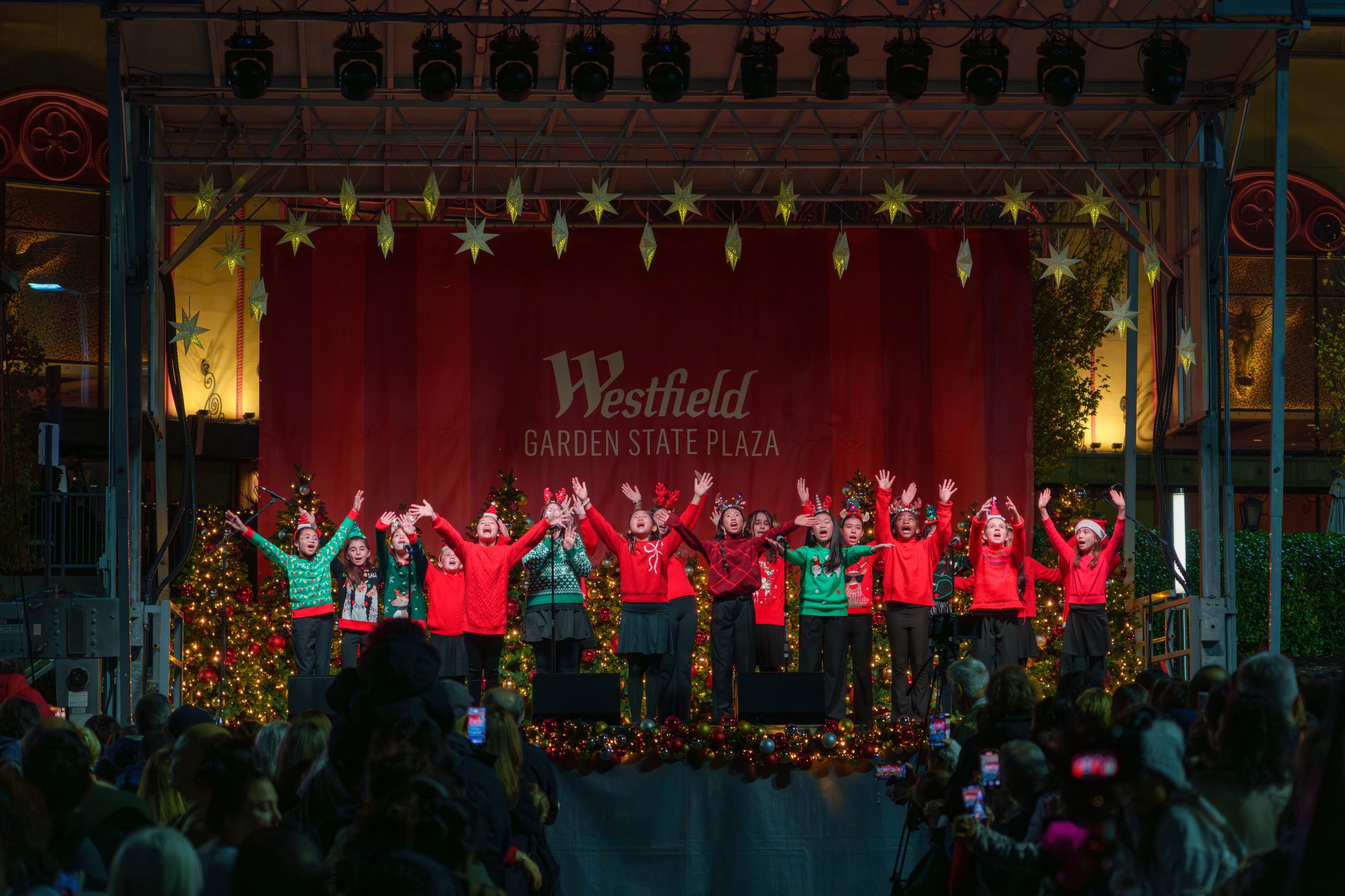 Children performing on stage at a Christmas event during the holiday season, with Christmas trees and festive decorations, on a stage labeled 'Westfield Garden State Plaza'.