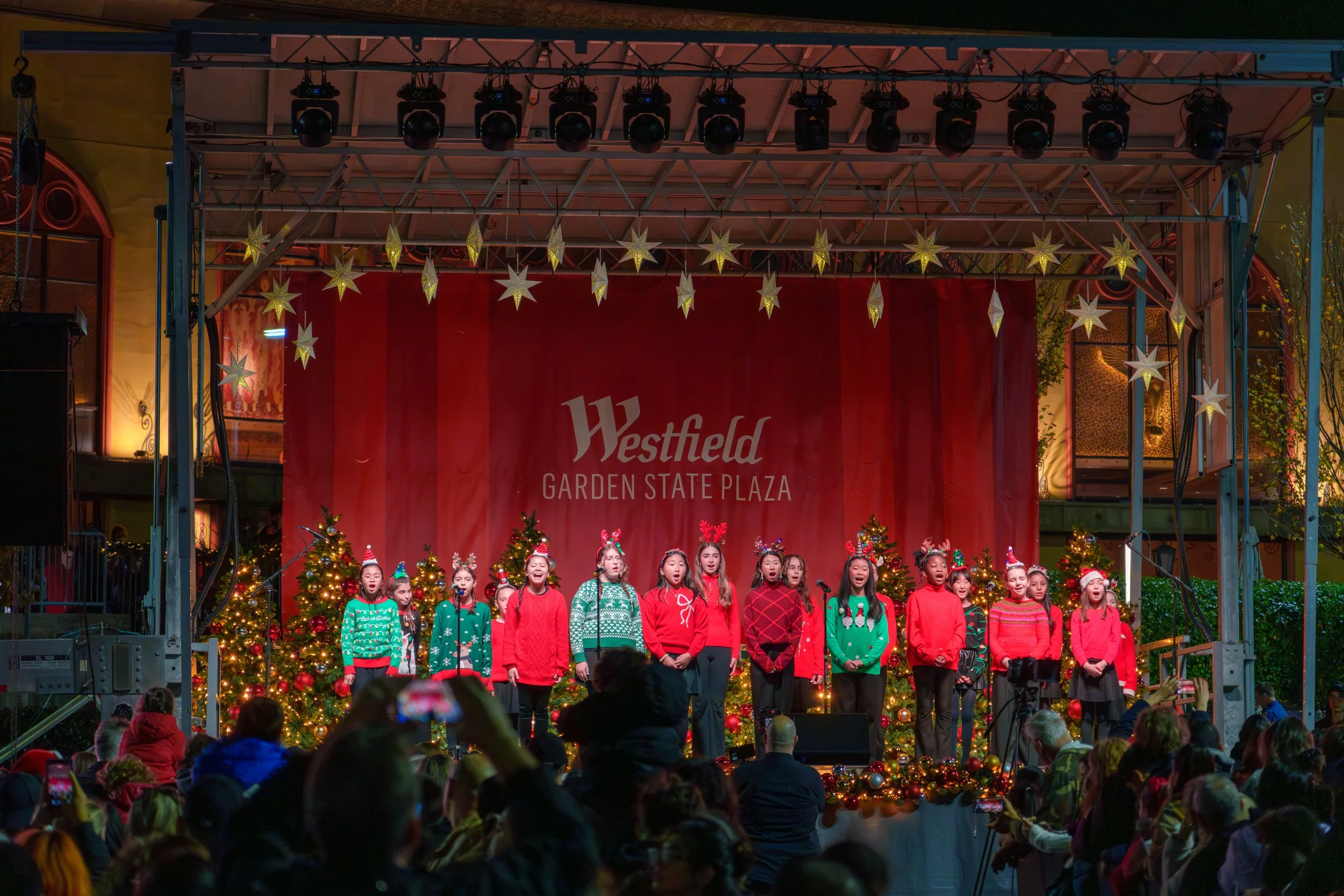 Children wearing festive holiday sweaters and accessories singing on stage decorated with Christmas trees at Westfield Garden State Plaza during a holiday performance, with an audience watching.