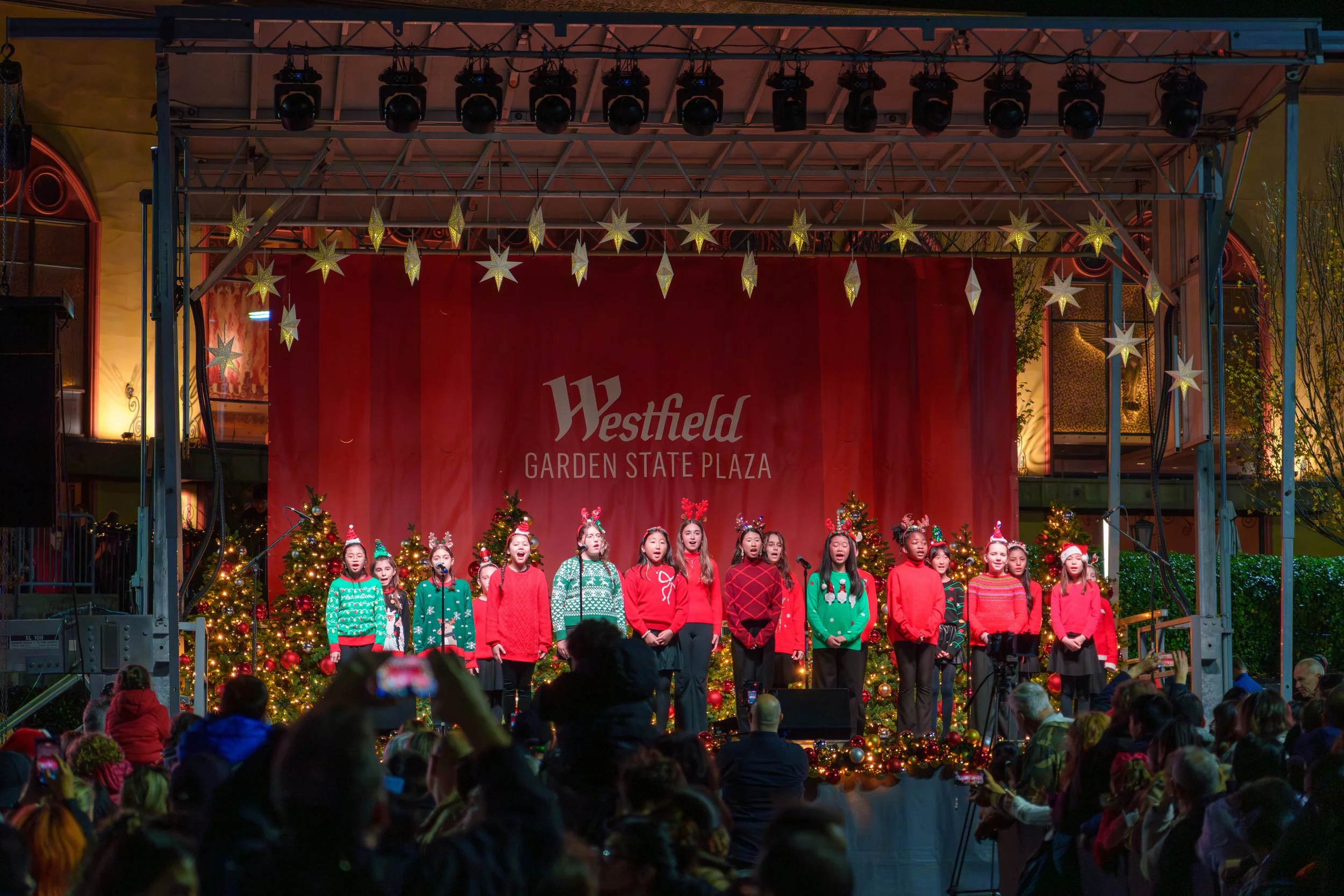 A group of children performing on stage at a Christmas event in Westfield Garden State Plaza, decorated with Christmas trees, lights, and star-shaped ornaments.