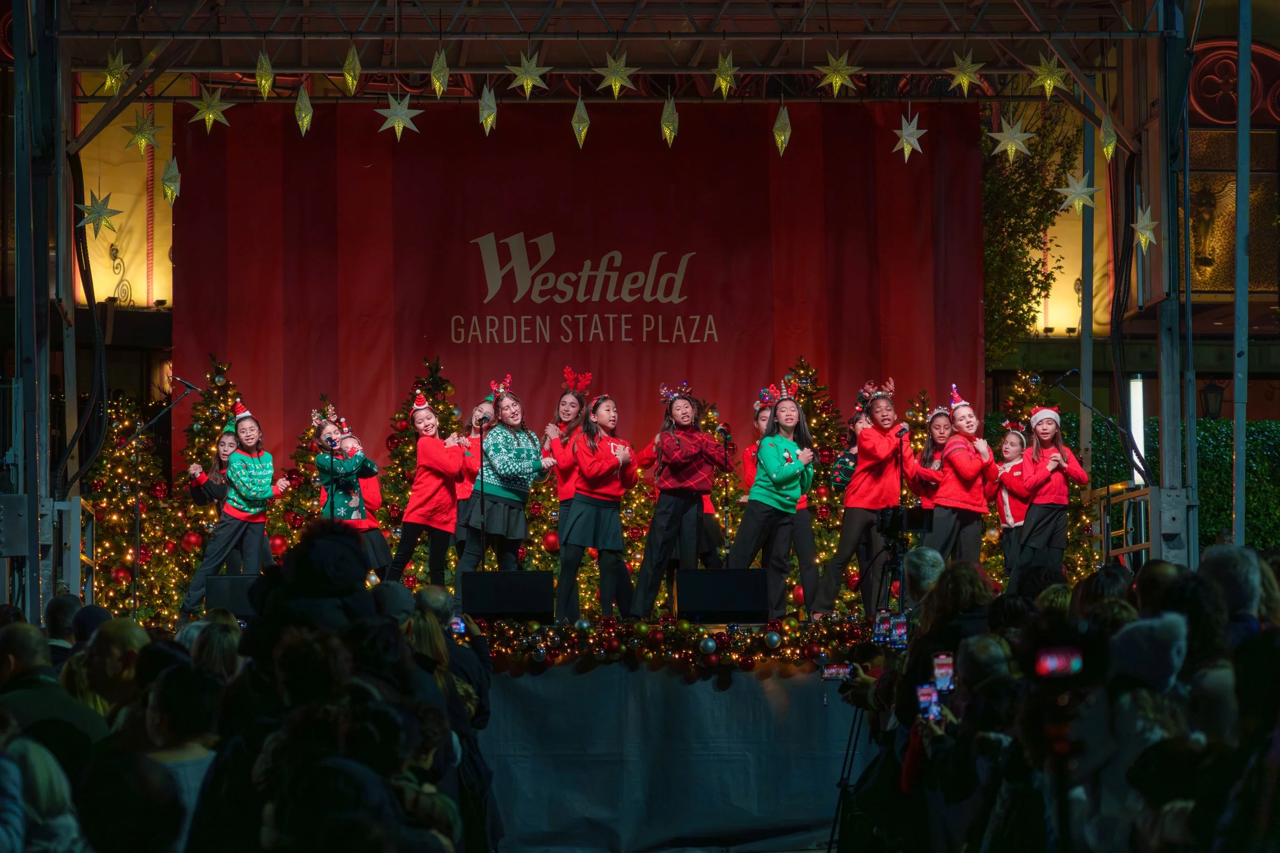 Children performing a Christmas show on stage at Westfield Garden State Plaza, decorated with Christmas trees, ornaments, and holiday lights, with an audience watching.