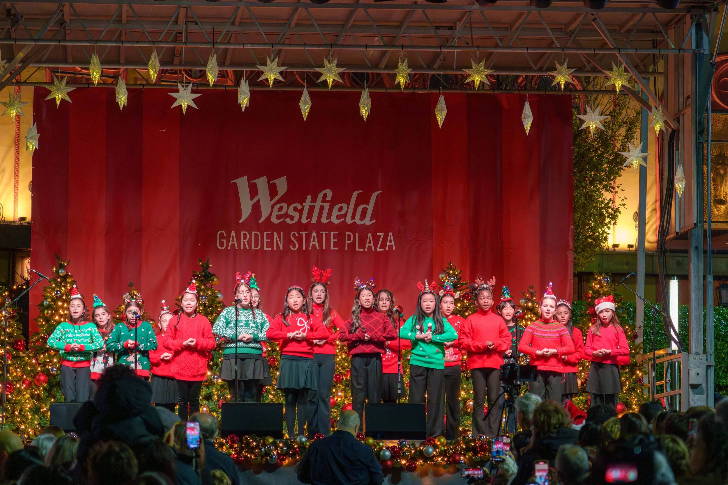 Children performing a Christmas concert on stage at Westfield Garden State Plaza, decorated with Christmas trees, ornaments, and festive lights.