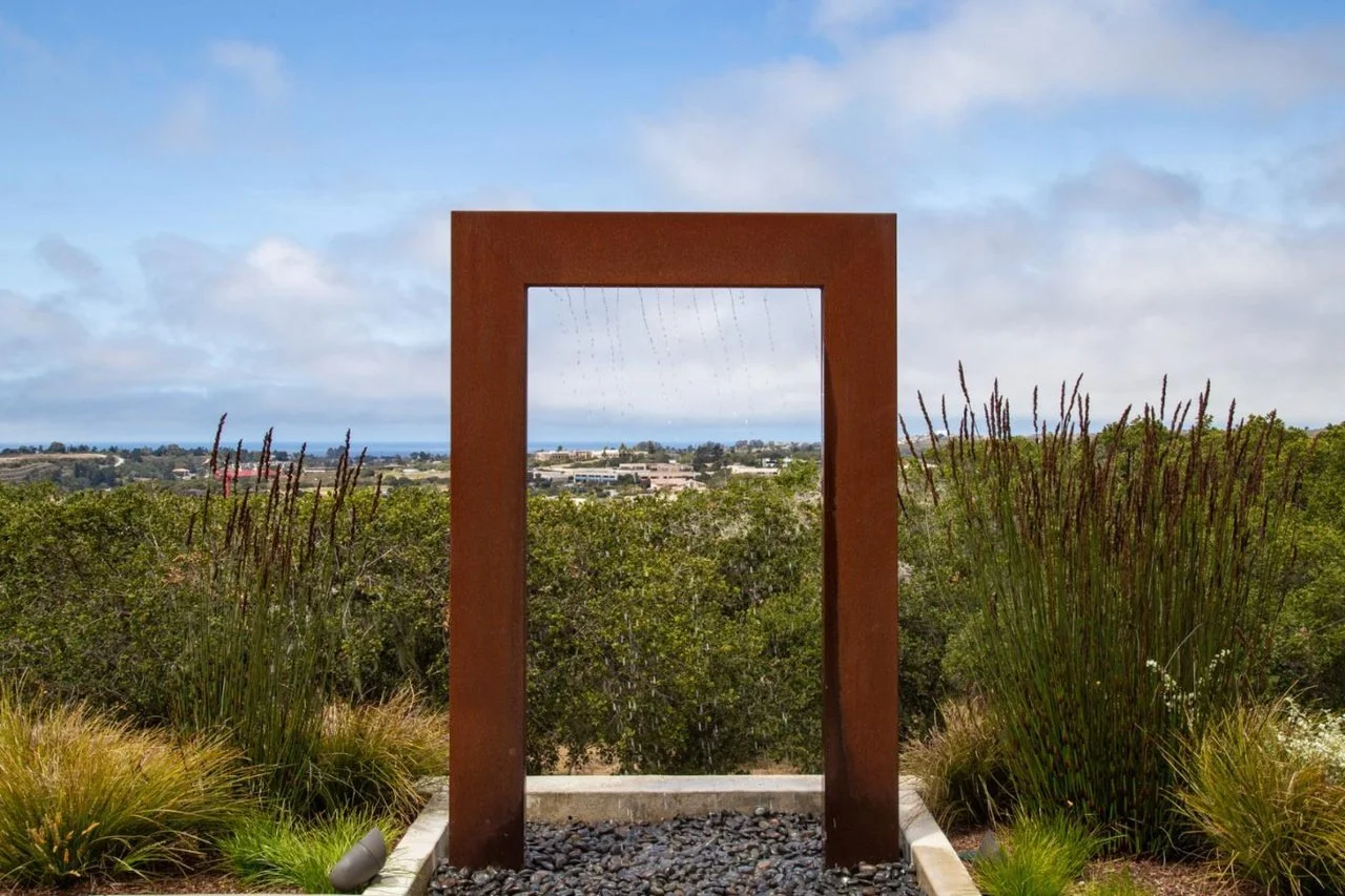 A rust-colored metal rectangular frame standing outdoors with a view through it of a landscape with trees, buildings, and a distant coastline under a cloudy sky.
