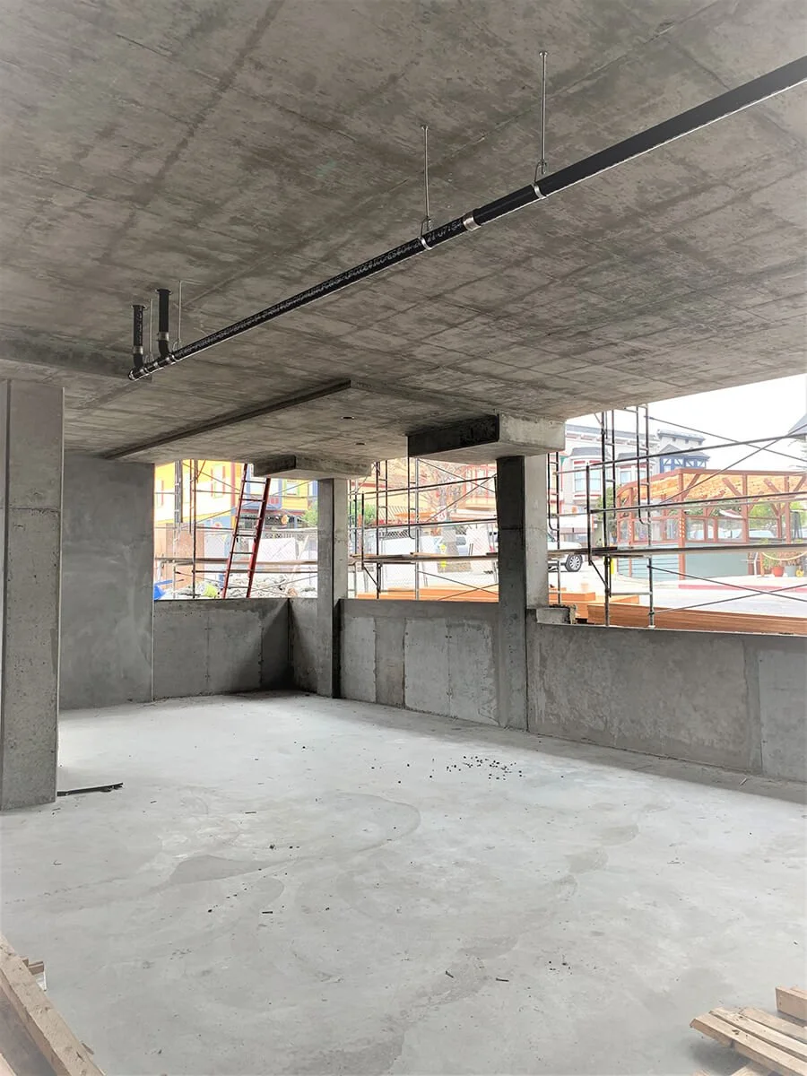 Construction site showing an unfinished room with concrete walls and ceiling, exposed plumbing pipe on the ceiling, and scaffolding outside the windows.