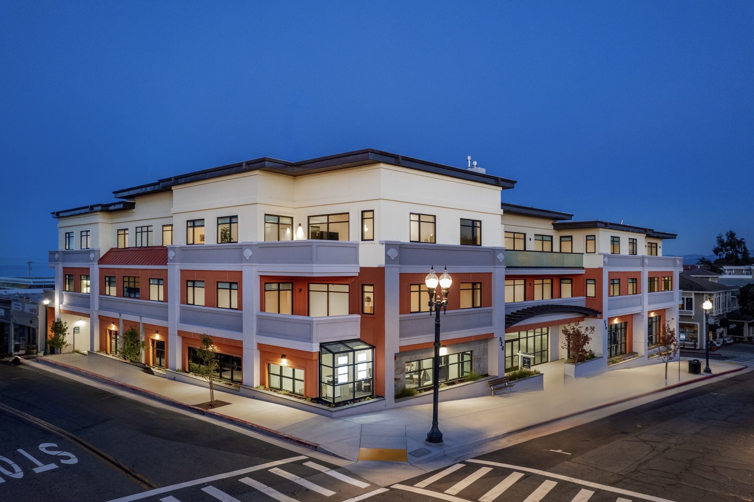 A modern multi-story building at dusk with exterior lighting, large windows, and a glass entrance, situated on a street with street lamps and a crosswalk.