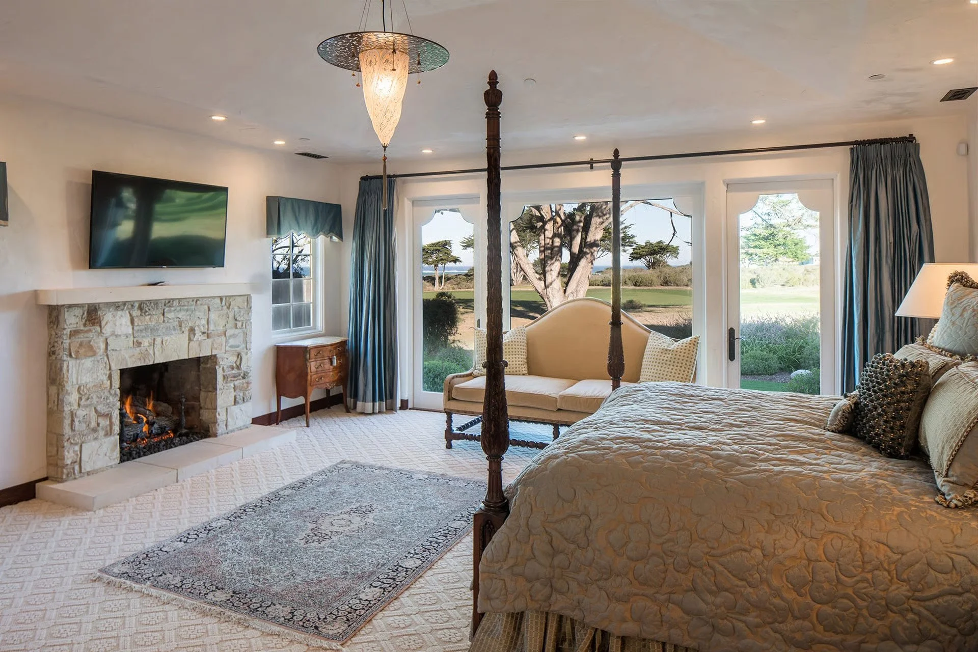 A bedroom with a four-poster bed, a fireplace with a television above it, a cream-colored sofa, a small wooden side table, and large windows showing a view of trees and grass outside.