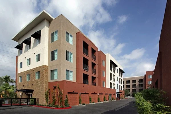 Multi-story apartment complex with colorful exterior walls, featuring beige, red, and white sections, with balconies and large windows, under a partly cloudy sky.