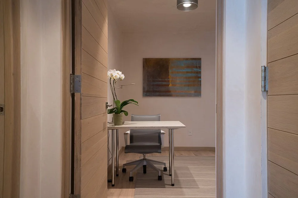 View of a small office or meeting room with a white table, black and gray office chairs, a potted orchid plant, a wall-mounted painting, and wooden panel walls.