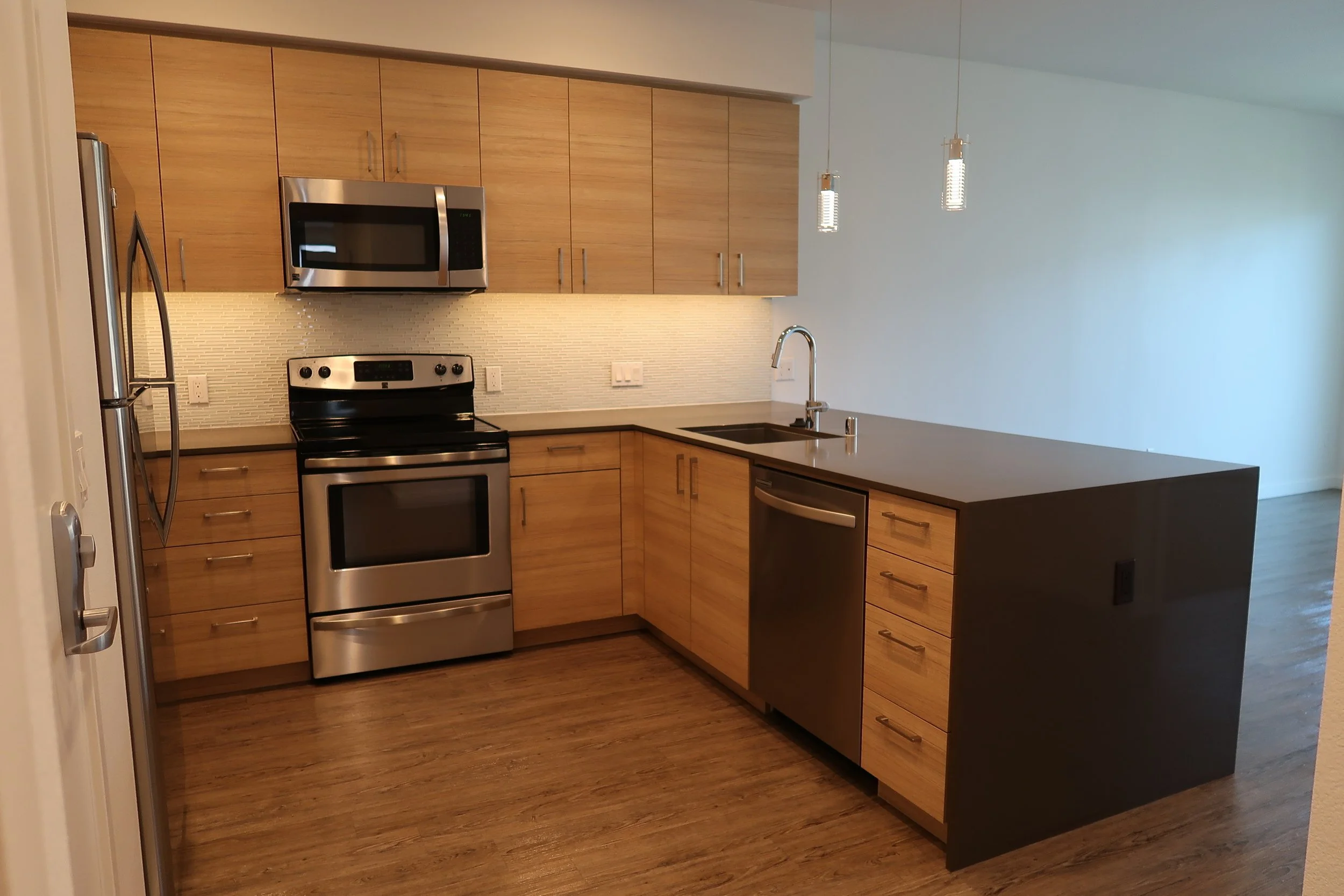 Modern kitchen with wooden cabinets, stainless steel appliances including a microwave and oven, black countertop with a sink, pendant lights, and a hardwood floor.