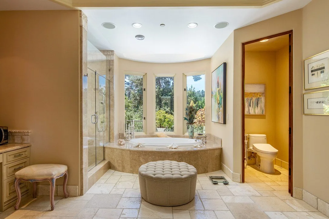 Luxurious bathroom with a large bay window, soaking tub, and a glass-enclosed shower. Light-colored tiled floor and beige walls. There is a small beige tufted ottoman and framed artwork.