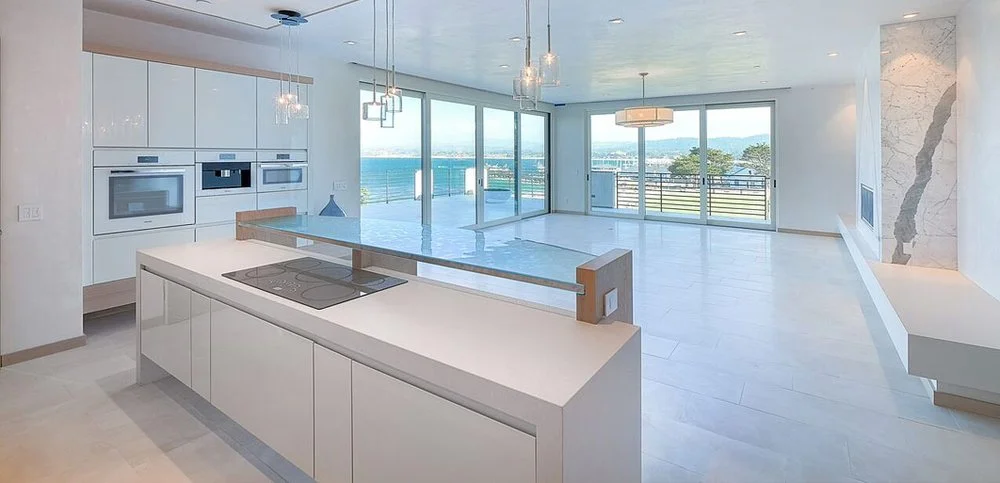 Bright, modern open-concept kitchen and living area with large glass windows overlooking a body of water and greenery, featuring white cabinetry, marble accents, and minimalist design.
