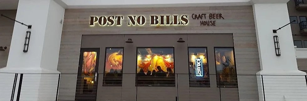 Exterior of a craft beer house named 'Post No Bills' with large windows and a neon sign inside. The building has white walls and black decorative lighting on either side.