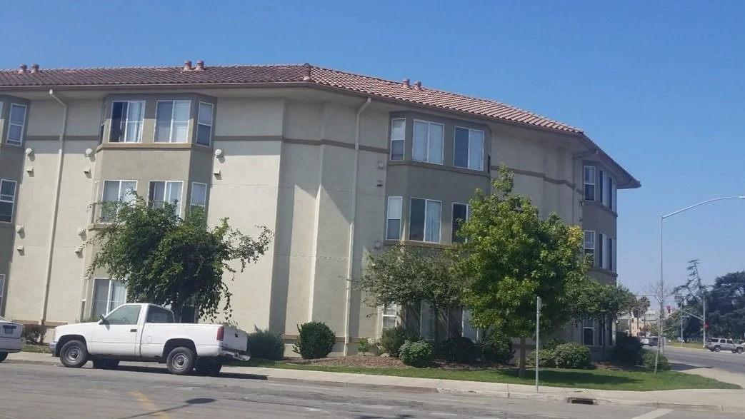 A beige, multi-story apartment building with a red tile roof, surrounded by trees and shrubs, with parked cars in front.