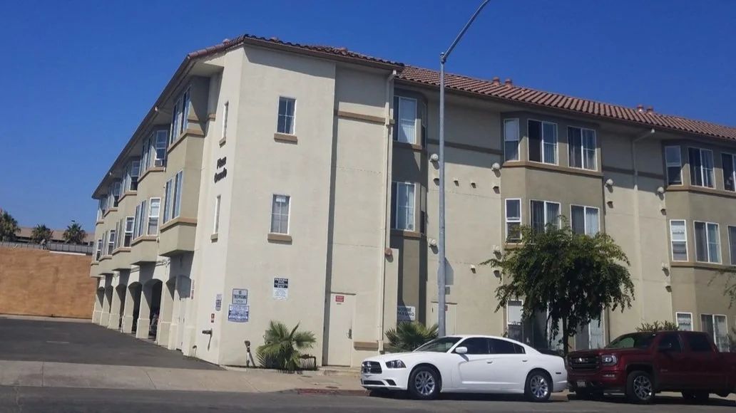 A beige multi-story apartment building with brown roof tiles, multiple windows, and parking spaces in front, with a white car in the foreground and a tree on the right side.