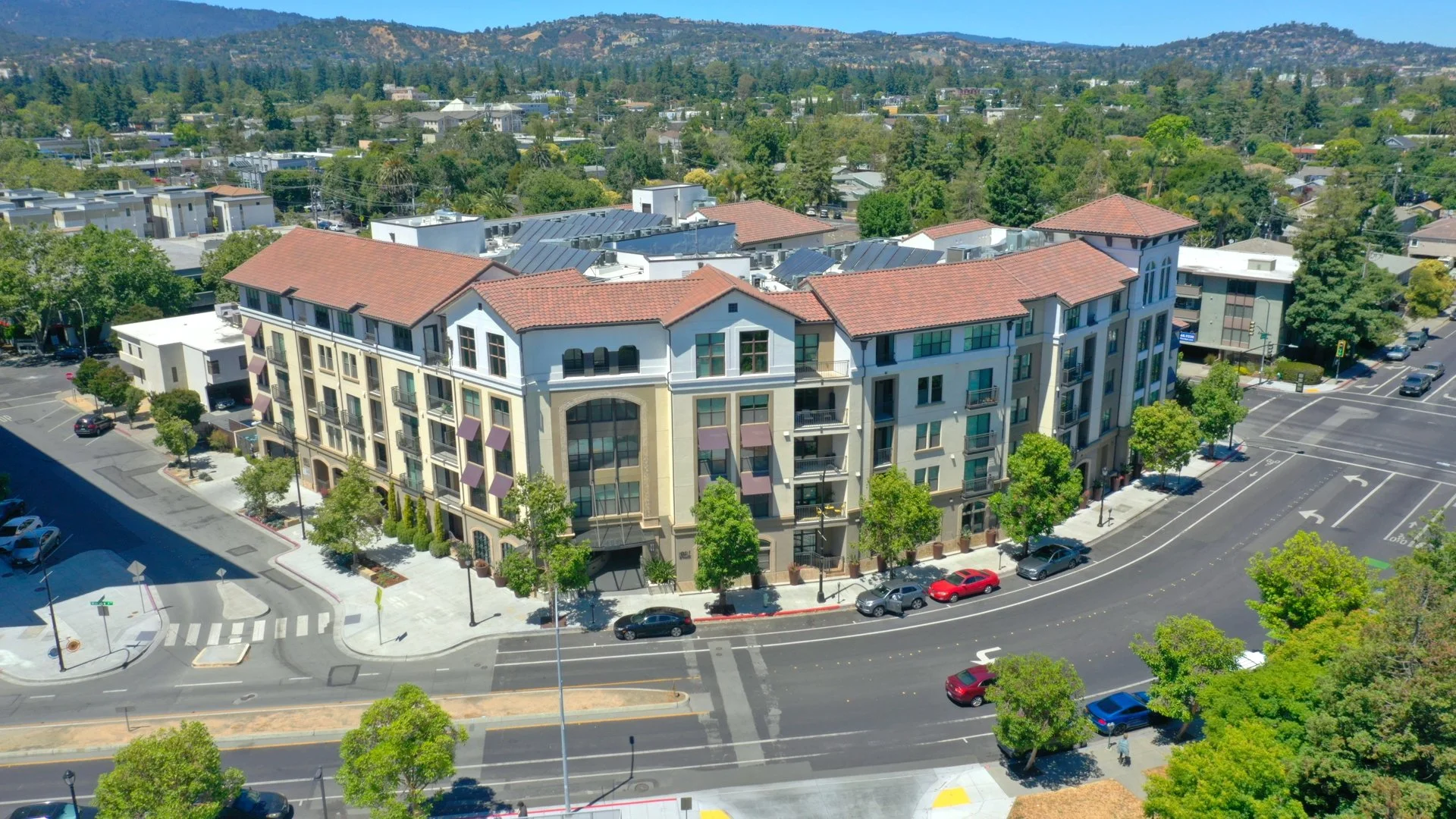 Aerial view of a multi-story residential building with terracotta roof tiles, surrounded by trees, parked cars, and nearby streets with crosswalks and traffic signals, in a suburban neighborhood with hills and forest in the background.