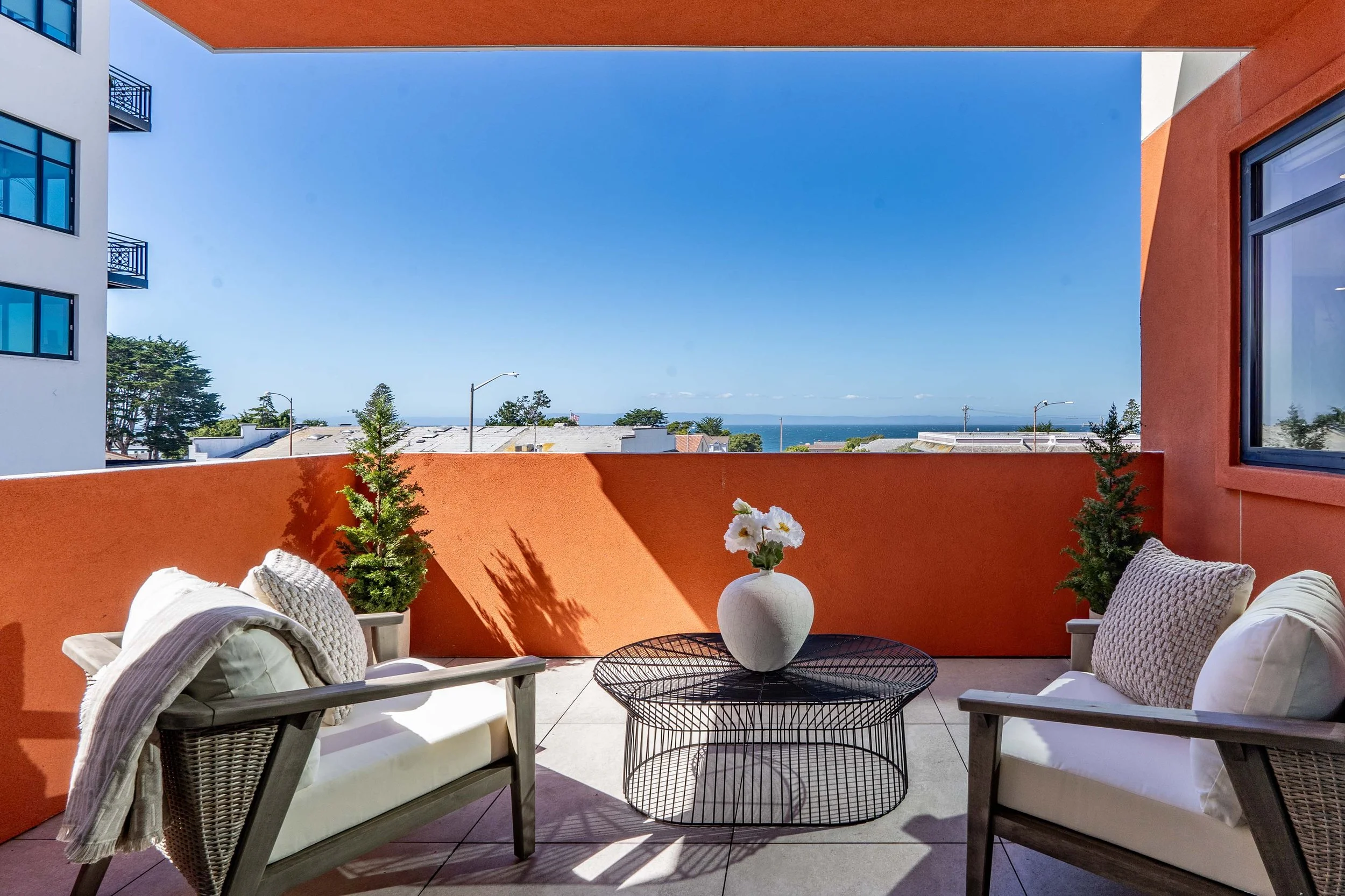Balcony with white cushioned chairs, a white vase with flowers, small potted plants, a black wire table, and an orange wall opening revealing a clear blue sky, trees, rooftops, and the ocean in the distance.