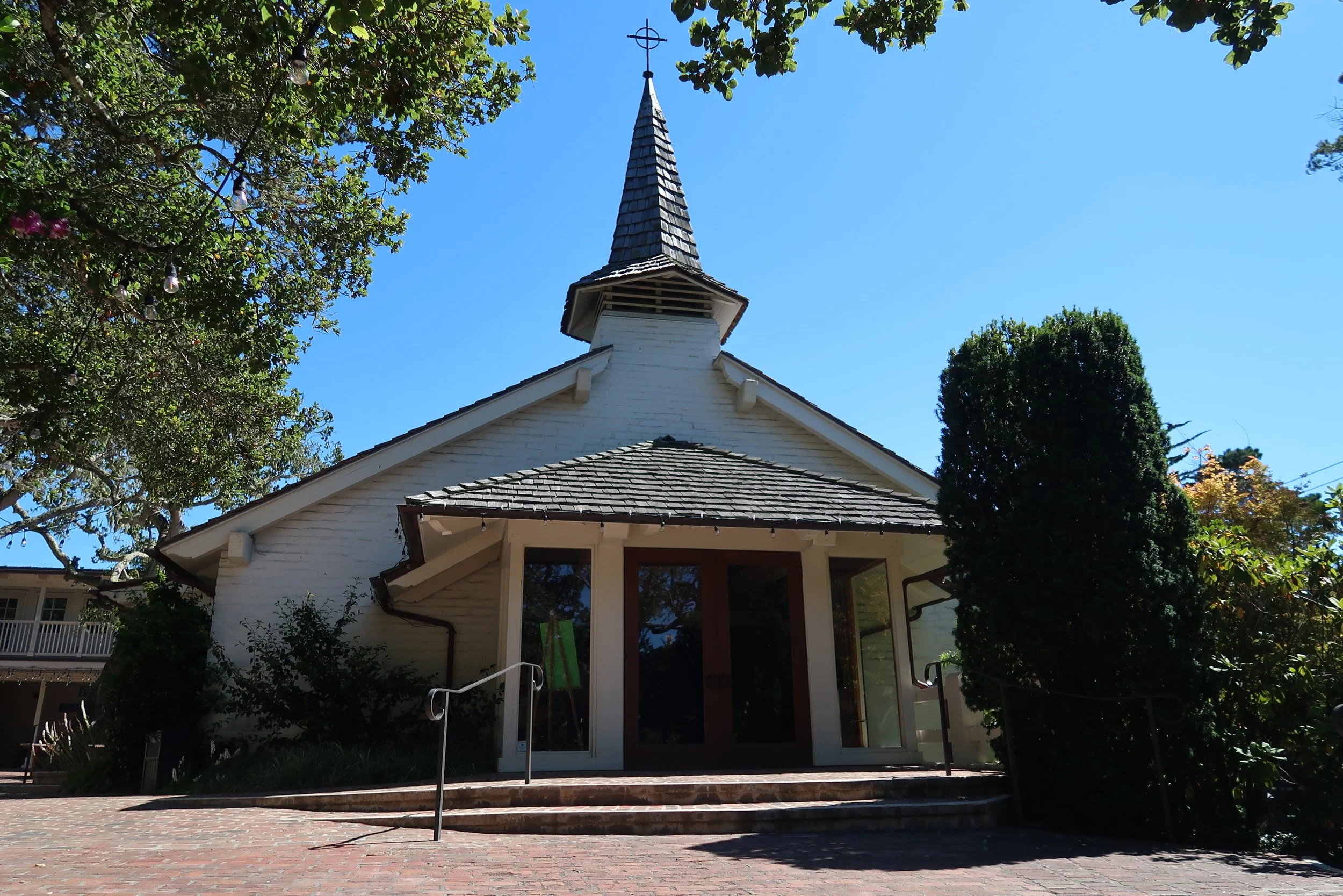 A white church building with a pointed steeple topped with a cross, surrounded by green trees under a clear blue sky.