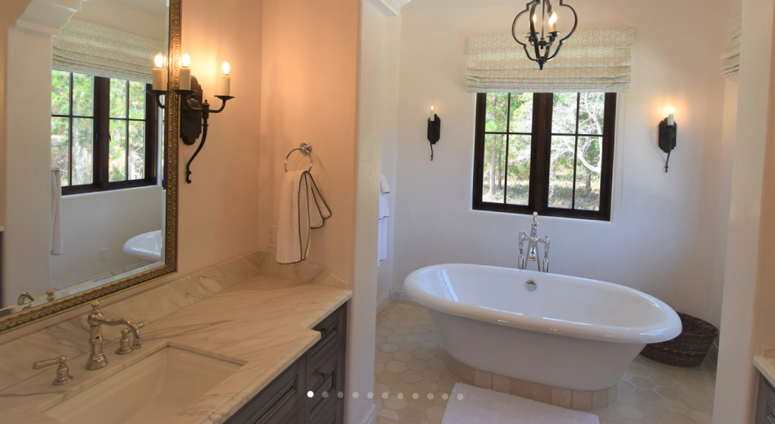 Bathroom with a freestanding bathtub by a window, a marble vanity with a mirror, and wall-mounted light fixtures in a light-colored room.