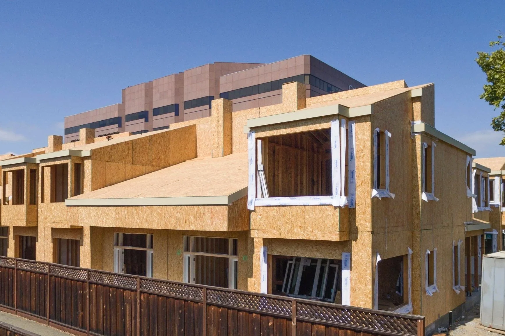 Under construction multi-story residential building with unfinished walls, windows, and exposed wooden framing, surrounded by a safety fence under a blue sky.