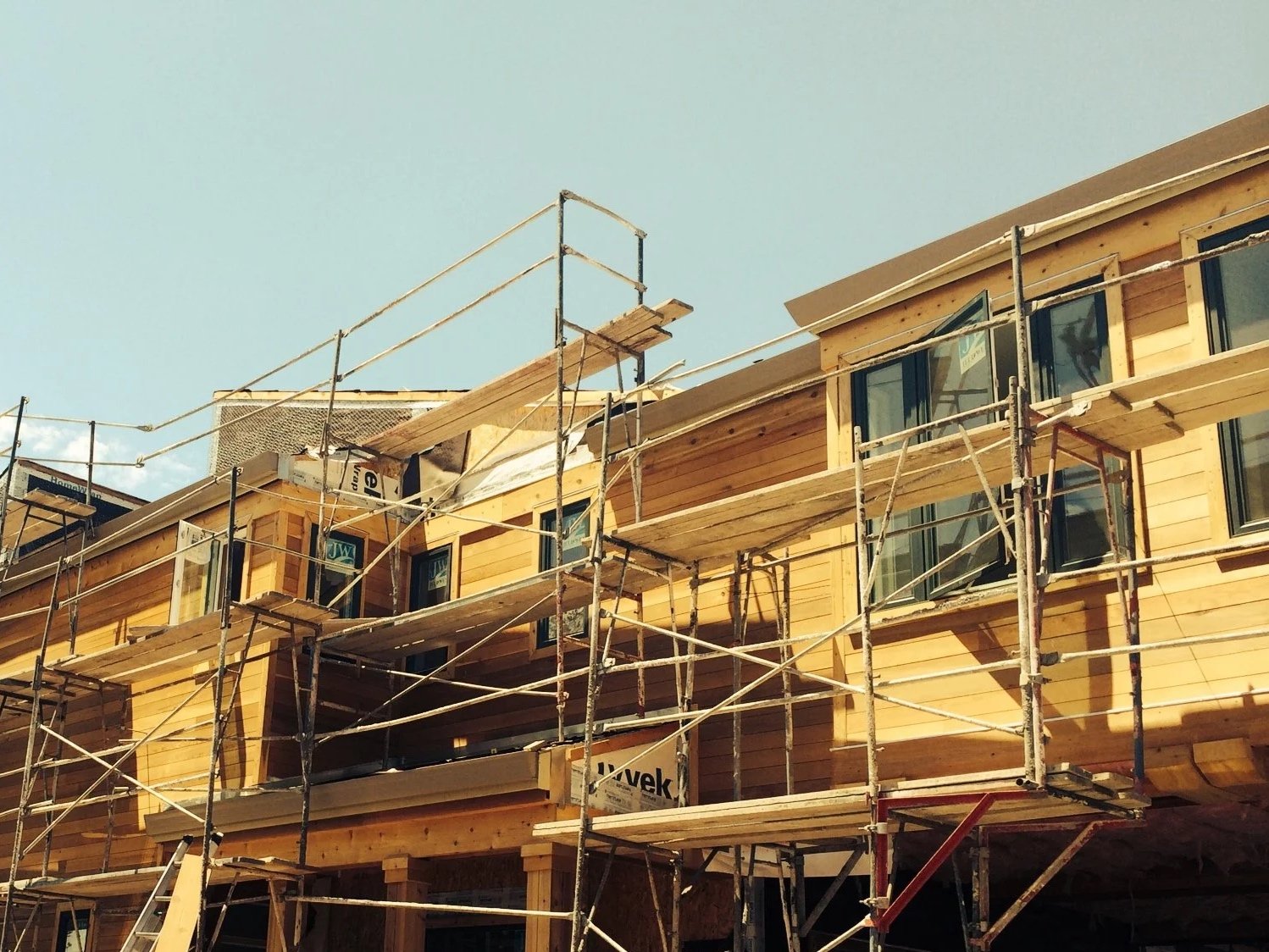 Wooden multi-story building under construction with scaffolding surrounding it, construction signs and a chain-link fence in the foreground, a tree on the left, and a clear blue sky above.