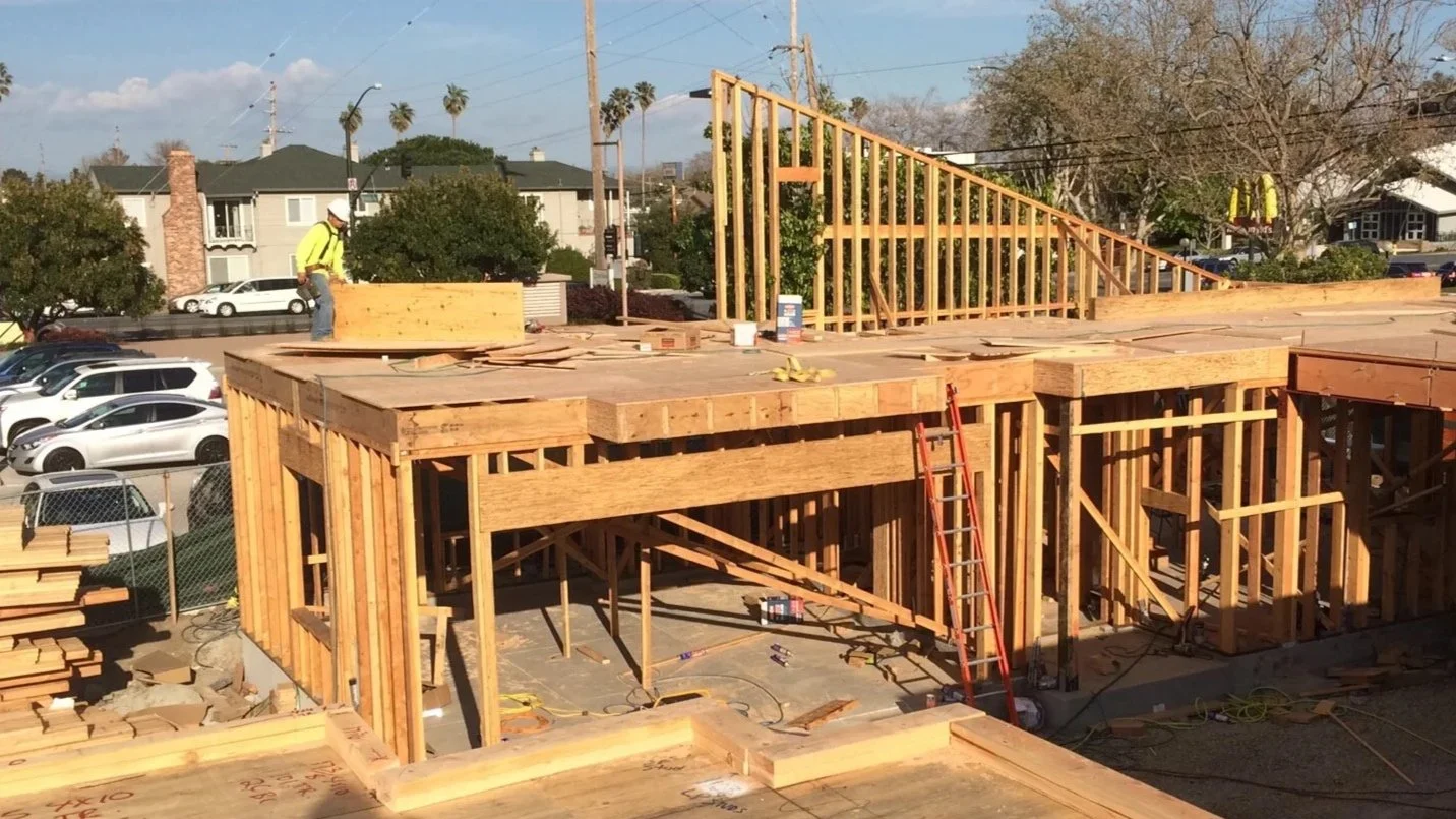 Construction site with wooden framing for a building, workers with safety helmets, and parked cars in the background.