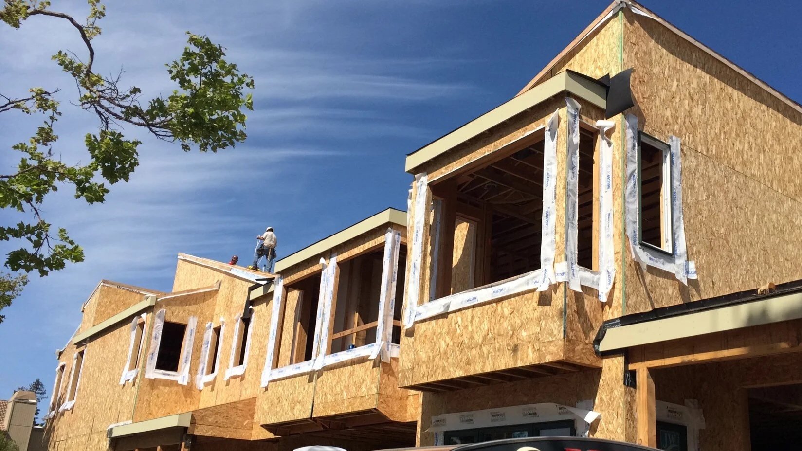 A multi-story residential building under construction with exposed wooden framework and sheathing. Two construction workers wearing hard hats work on the roof. A tree with green leaves is on the left side, and a bright blue sky is in the background.