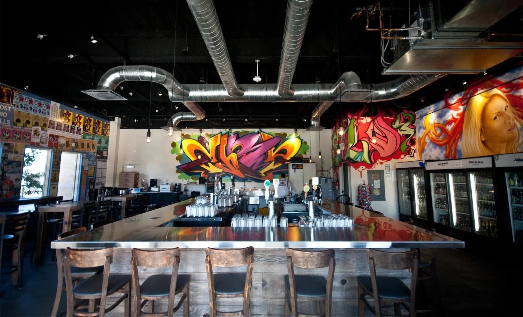 Interior of a modern bar or brewery with a large central stainless steel bar counter, graffiti-style colorful artwork on the wall, and painted murals including a woman's face. Industrial ceiling with exposed ducts and hanging lights, and refrigeratio