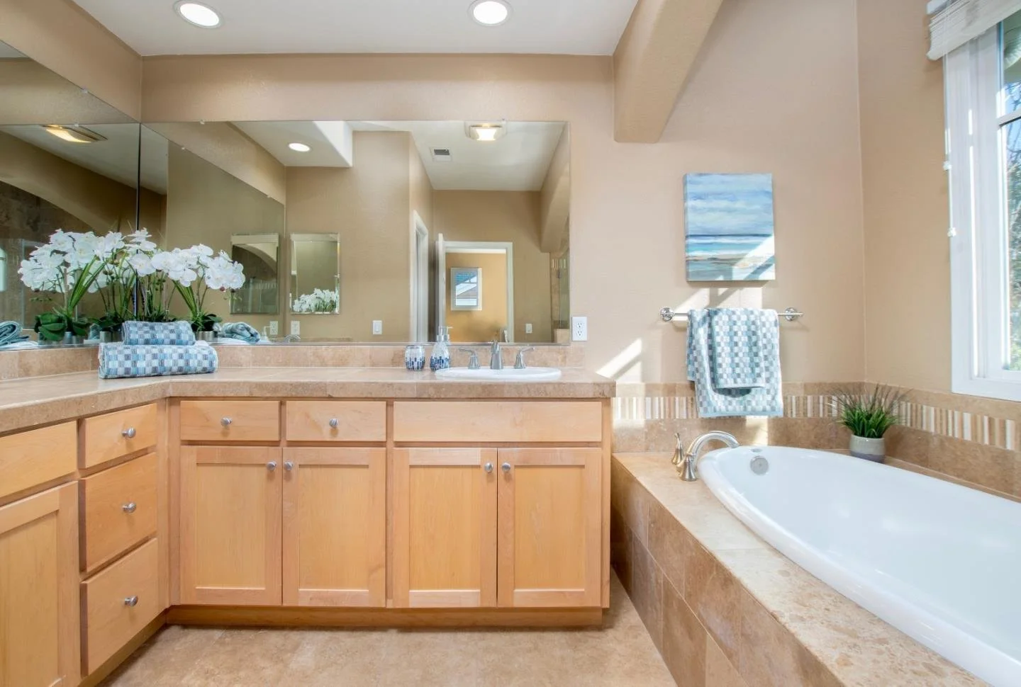 Bathroom with beige walls and cabinetry, large mirror, bathtub, decorative flowers, and blue towels.