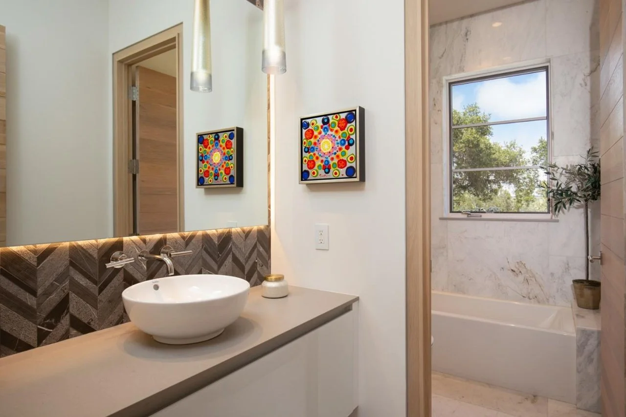 Modern bathroom with a white vessel sink on a beige countertop, framed vibrant artwork, a bathtub near a window with marble walls, and a potted plant.