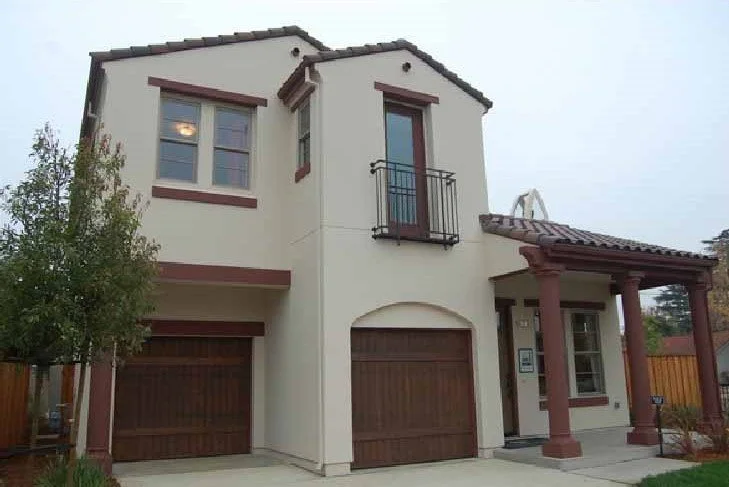 Two-story house with a beige exterior, brown trim, and a small front porch supported by two columns. There are two garage doors on the left side and a balcony with a black railing above the front door.