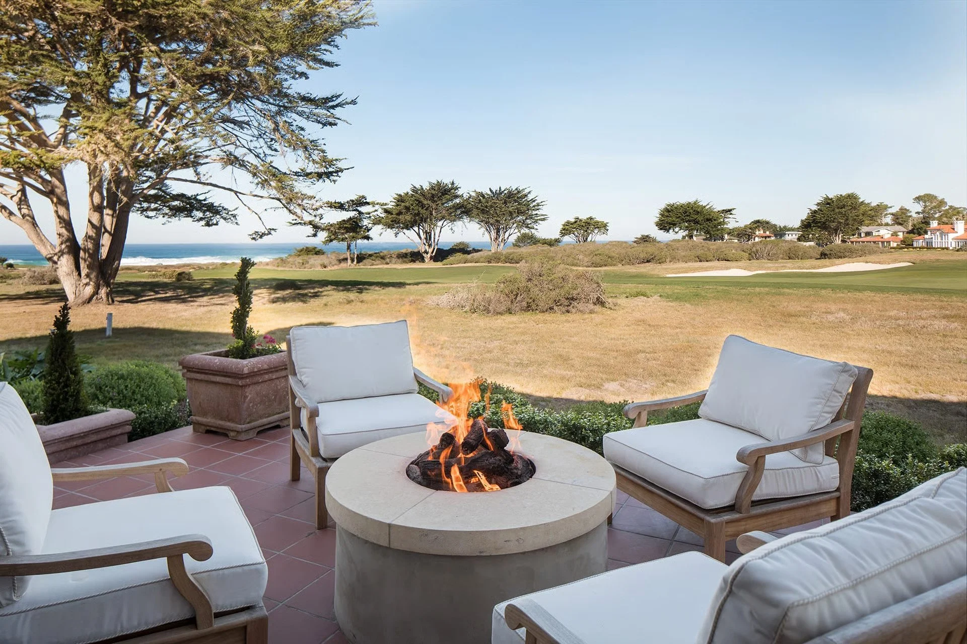 Outdoor patio with white cushioned chairs around a fire pit, overlooking a grassy area with trees and the ocean in the background.