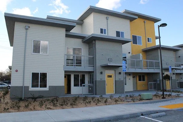 Exterior view of modern apartment building with a combination of white, gray, and yellow walls, multiple windows, and balconies, located in a residential area with a sidewalk, streetlamp, and designated handicapped parking signs.