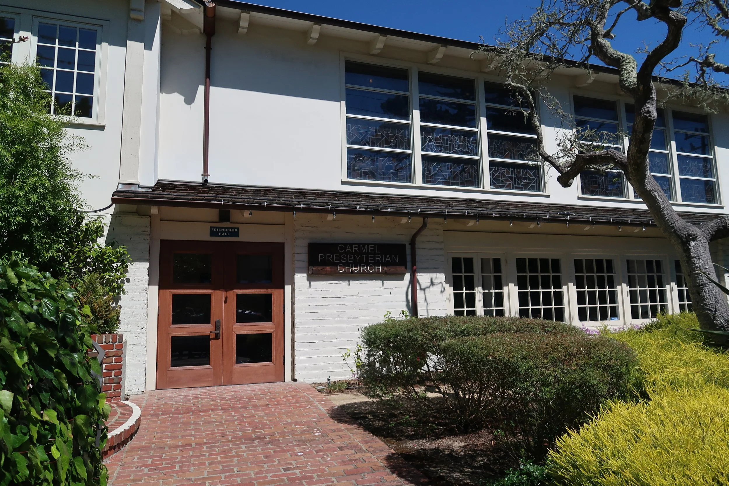 Exterior view of a white building with a sign reading 'Carmel Presbyterian Church' and 'Friendship Hall' above the door. There are large windows with stained glass, a brick pathway, and greenery including a leafless tree and bushes.