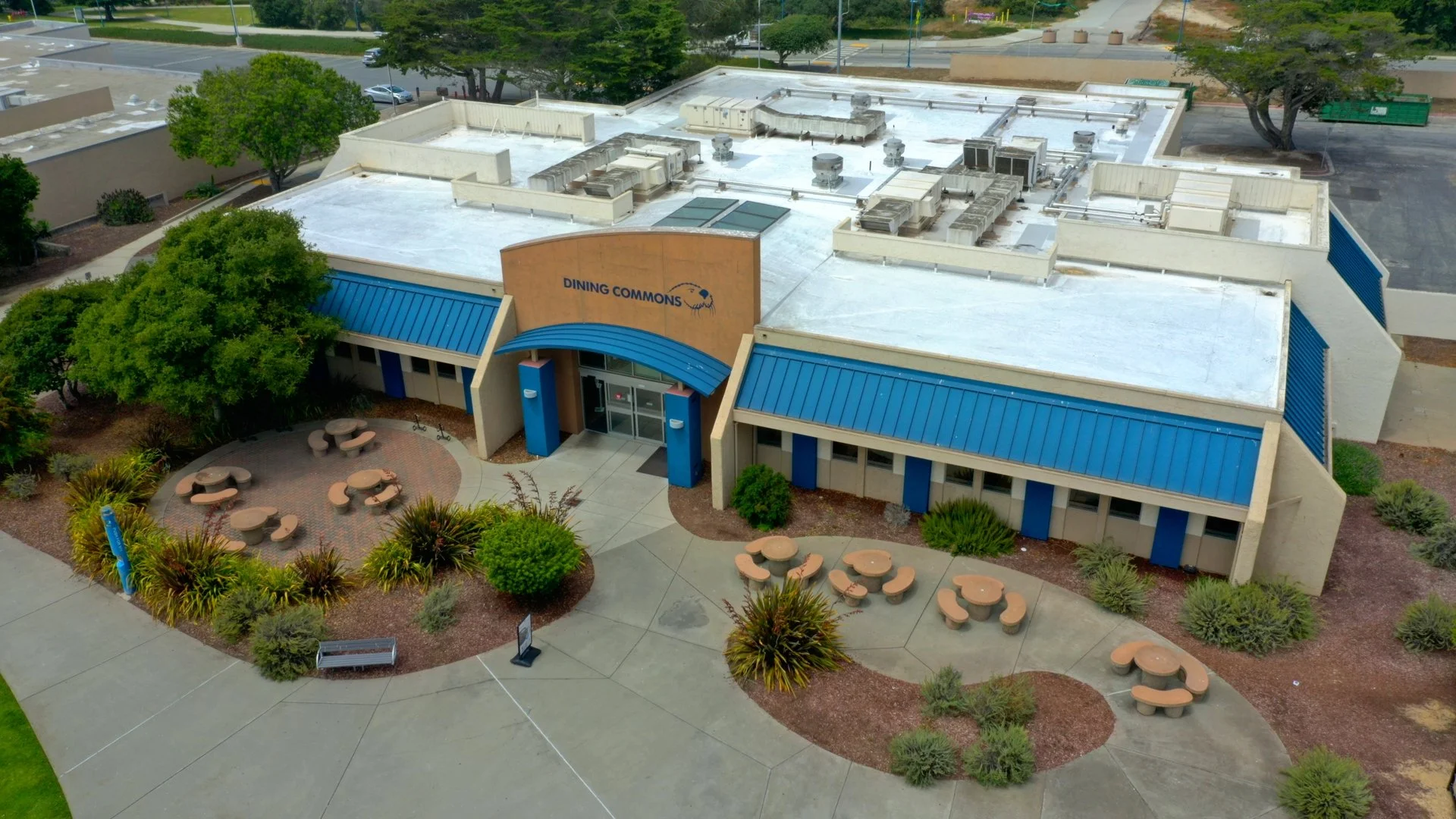 An aerial view of a building with a sign that says 'Dining Commons'. The building has a blue roof and beige walls, with a surrounding outdoor area featuring round tables and chairs, trees, and landscaped bushes.