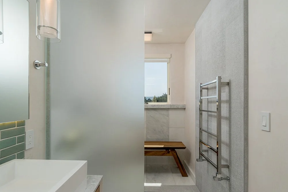 Bathroom with frosted glass partition, wall-mounted mirror, sink, window, wooden bench, and wall-mounted towel rack.