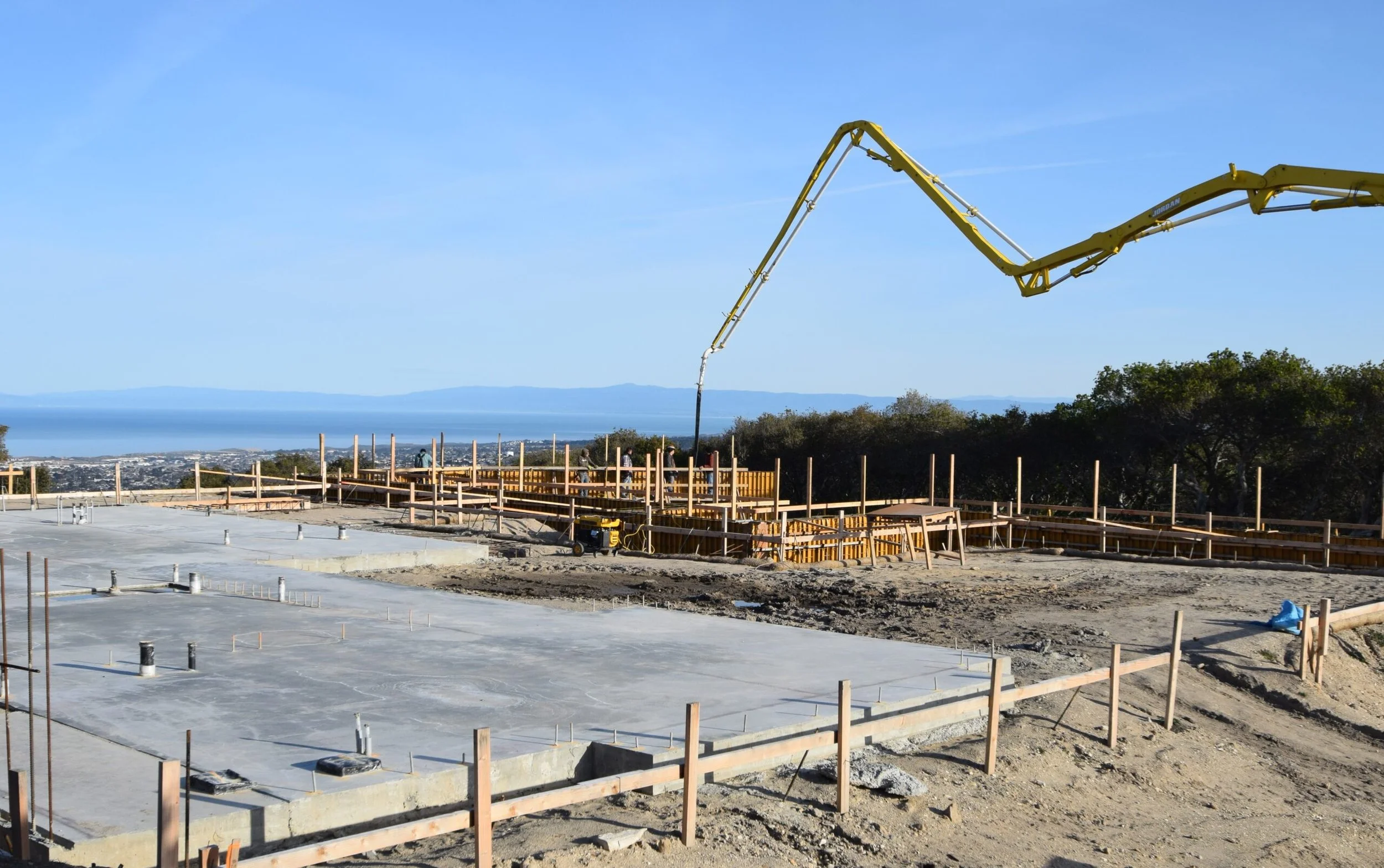 Construction site with concrete foundation, wooden framing, and a large boom pump. Clear blue sky and distant landscape in background.