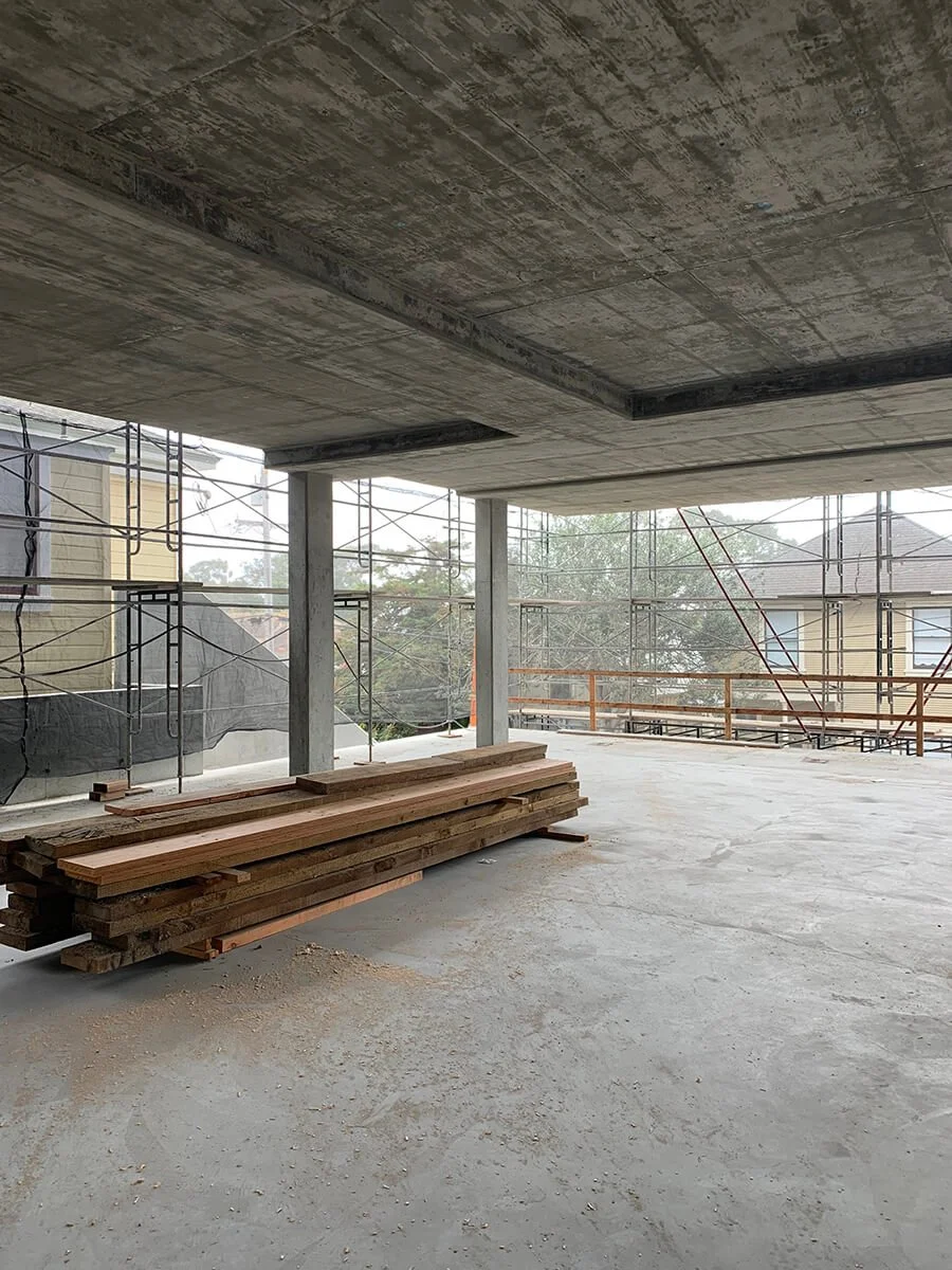 Construction site with a concrete ceiling, metal scaffolding, and a stack of wooden planks on the unfinished concrete floor.