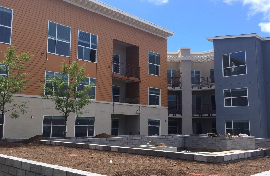 Construction site of a multi-story residential building with orange and gray exterior walls, large windows, and balconies, with newly planted trees and unfinished landscaping.