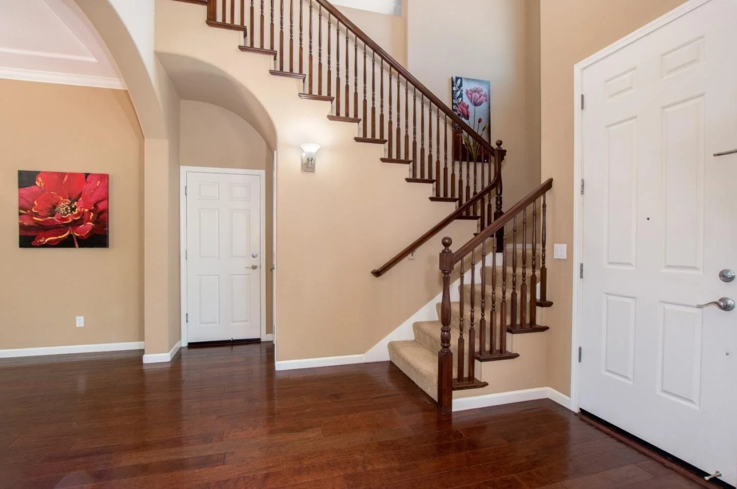 Interior of a house entryway showing a staircase with wooden banister and carpeted steps, a white front door, beige walls, and artwork on the walls.